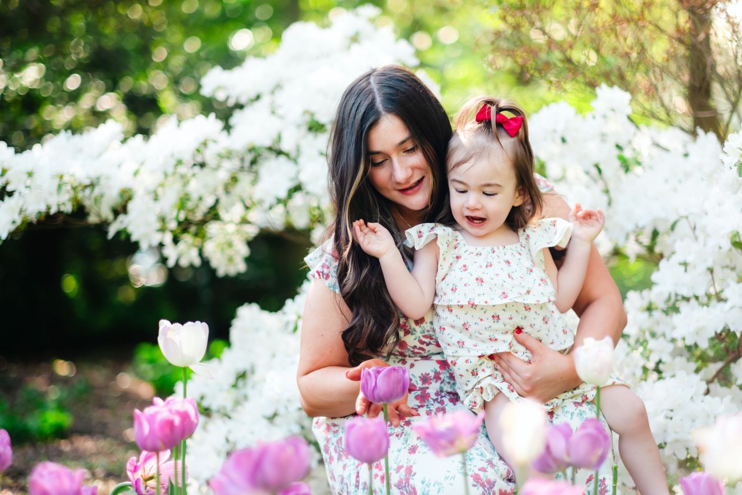 Mother holding young daughter in floral dresses among blooming white flowers and pink tulips in spring garden.