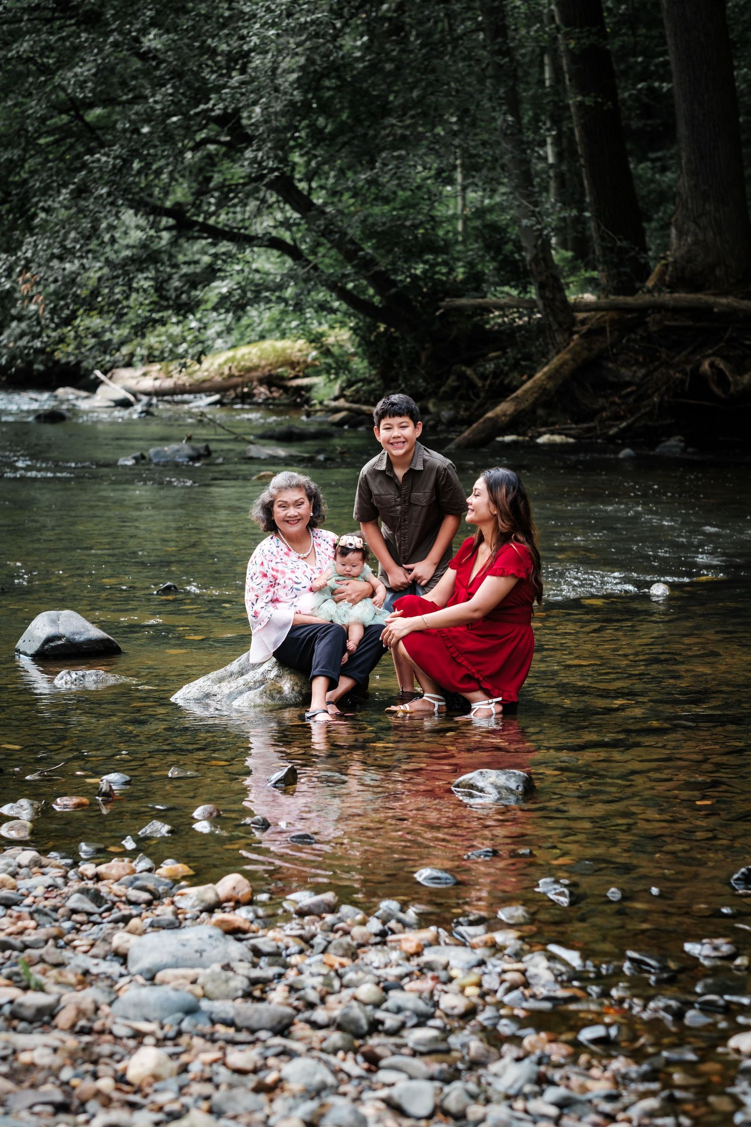 Family of four posing together in a rocky shallow river surrounded by lush green forest trees.