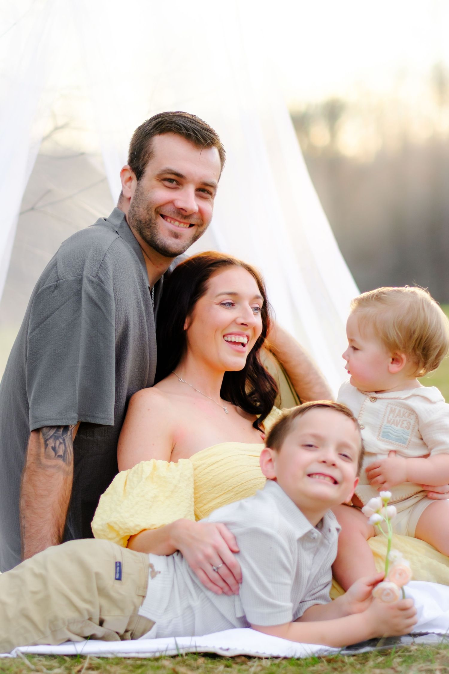 Happy family of four posing outdoors on a blanket near a white tent, parents smiling with two young children.