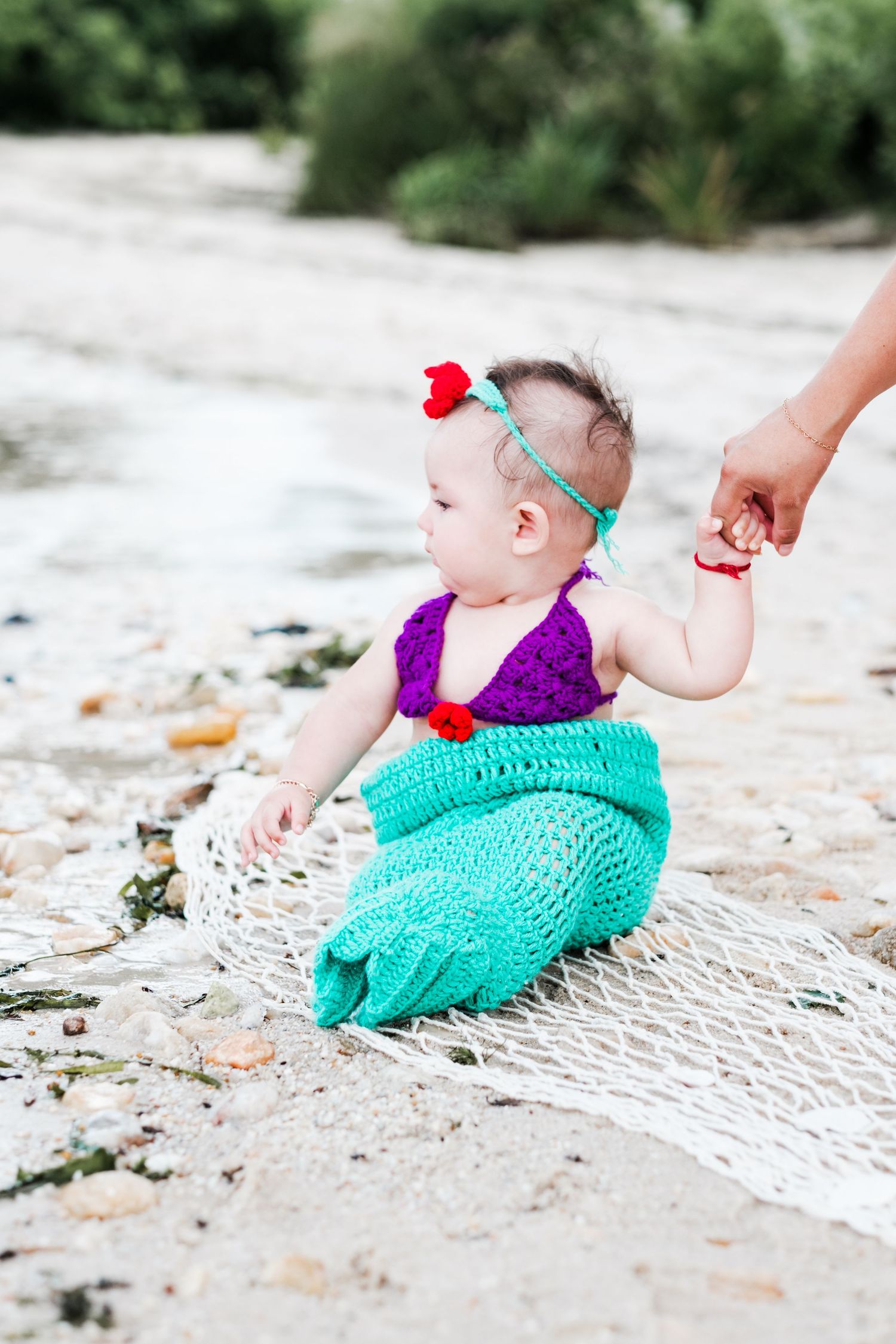 Baby girl in crochet mermaid costume with purple top and teal tail sitting on beach with fishing net.