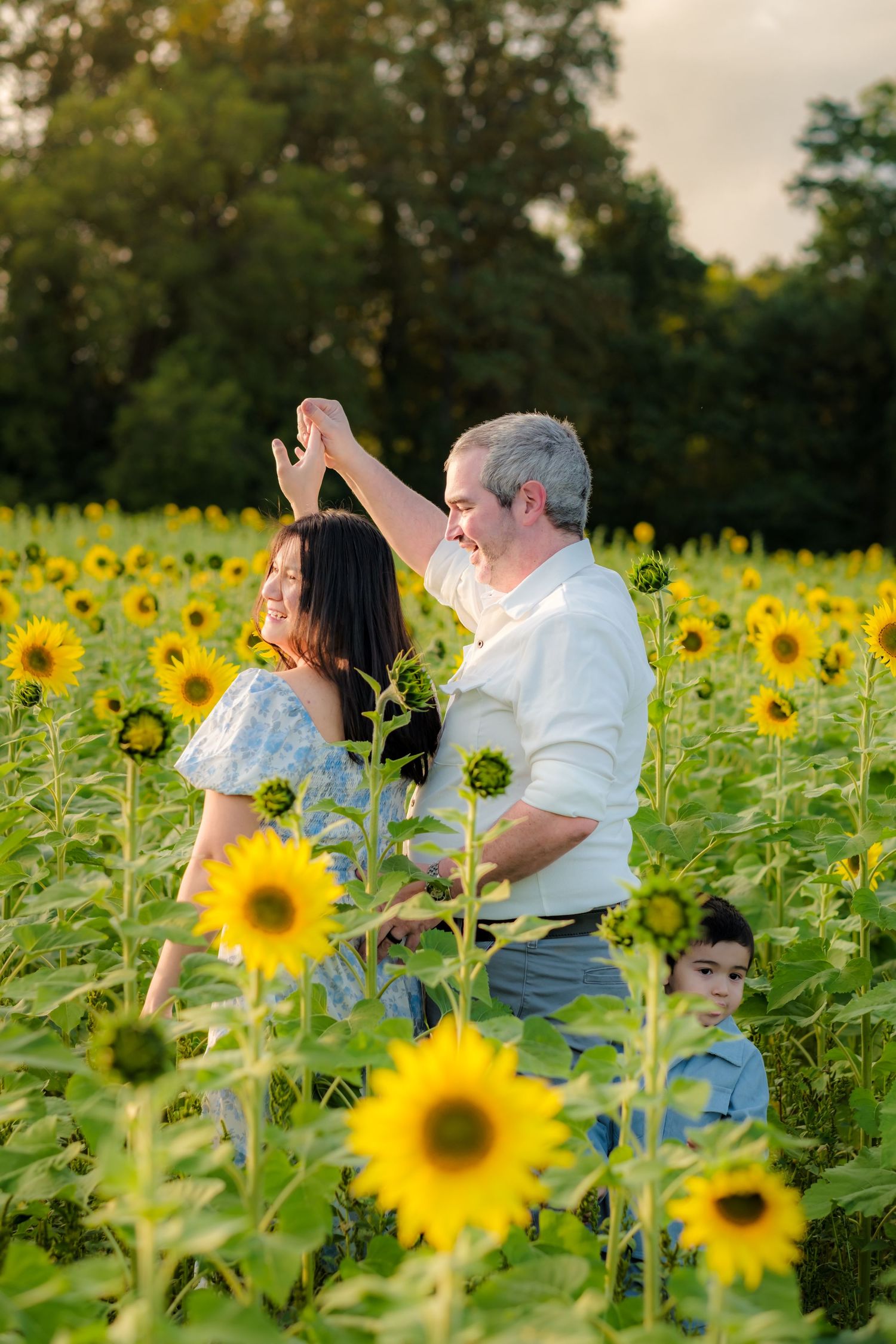 A couple dances joyfully in a sunflower field at golden hour, with a young boy peeking through the blooms.