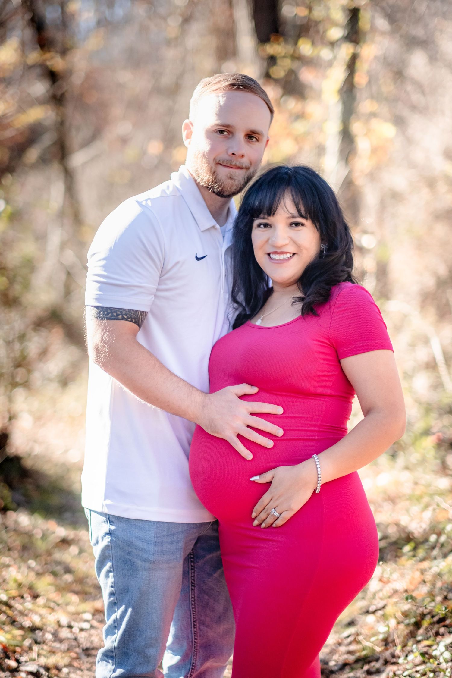 Happy couple posing outdoors for a maternity photo, woman in red dress cradling baby bump, man in white polo embracing her.