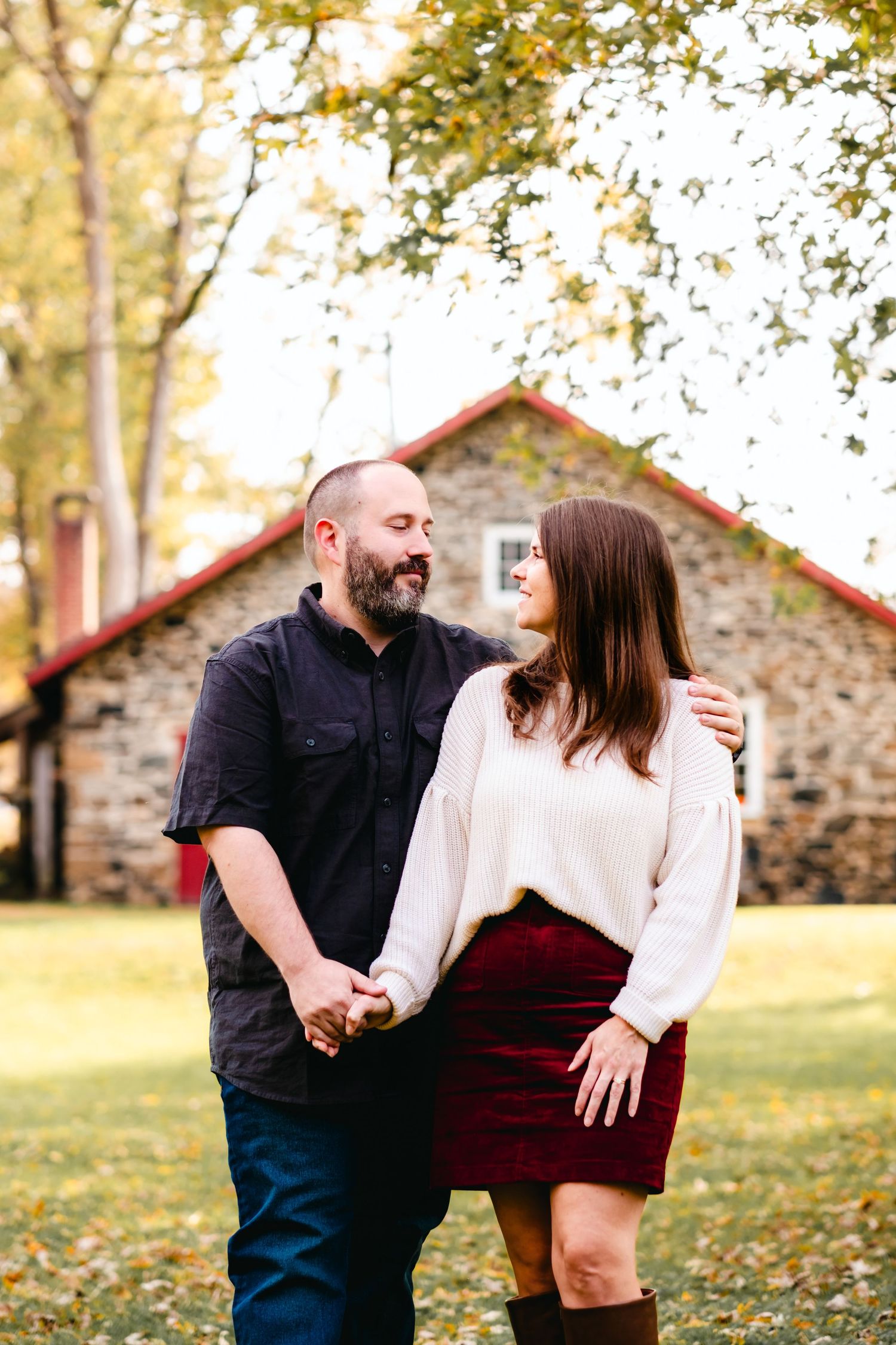 A couple holds hands and smiles at each other outdoors in autumn, with a stone building and colorful trees behind them.