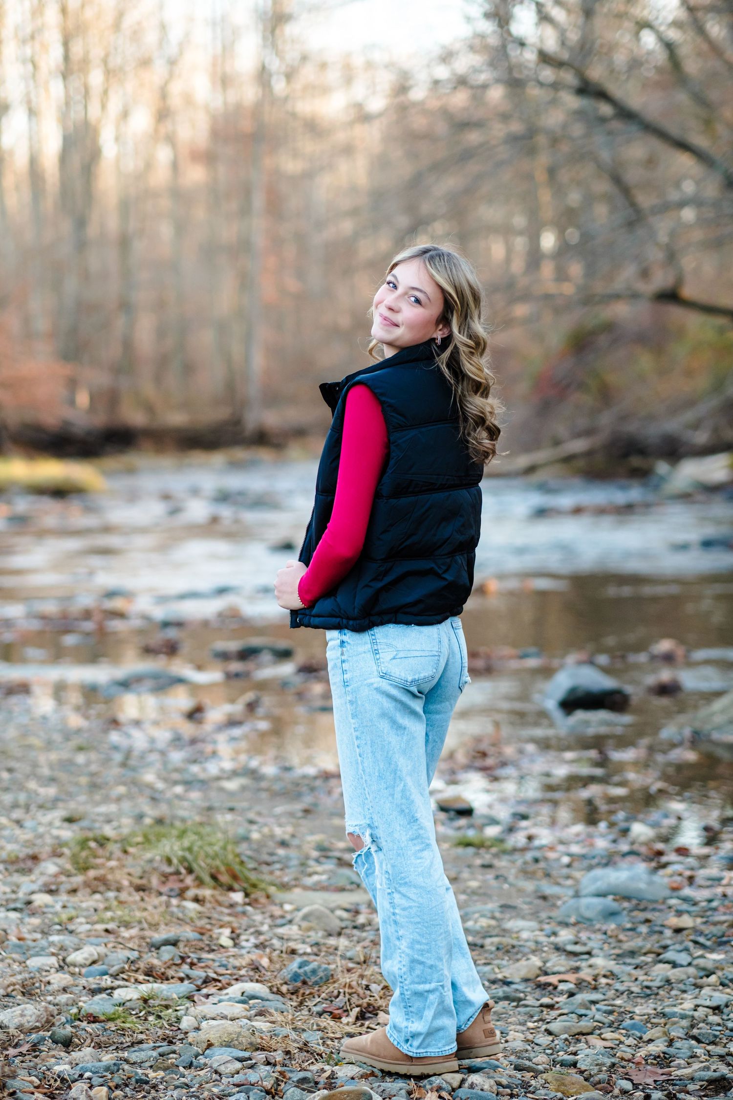 Teen girl in black puffer vest, red top, and ripped jeans standing by a rocky creek in autumn woods.