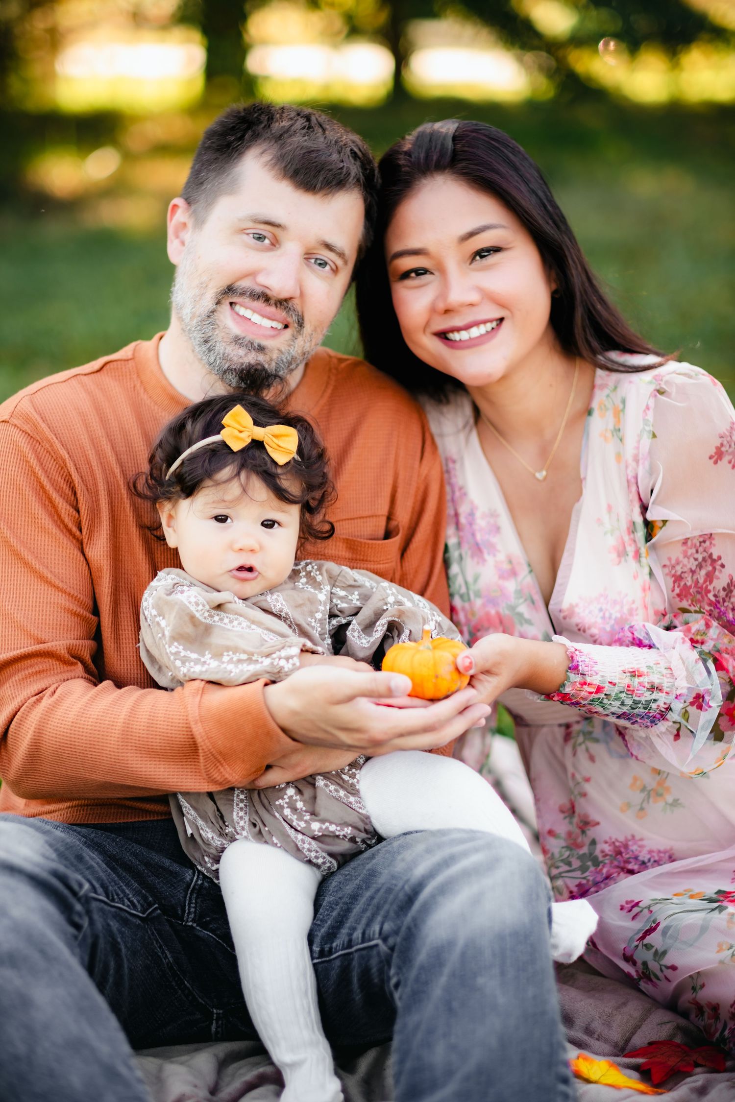 Happy family portrait with parents and baby girl holding a small pumpkin outdoors in a park setting.