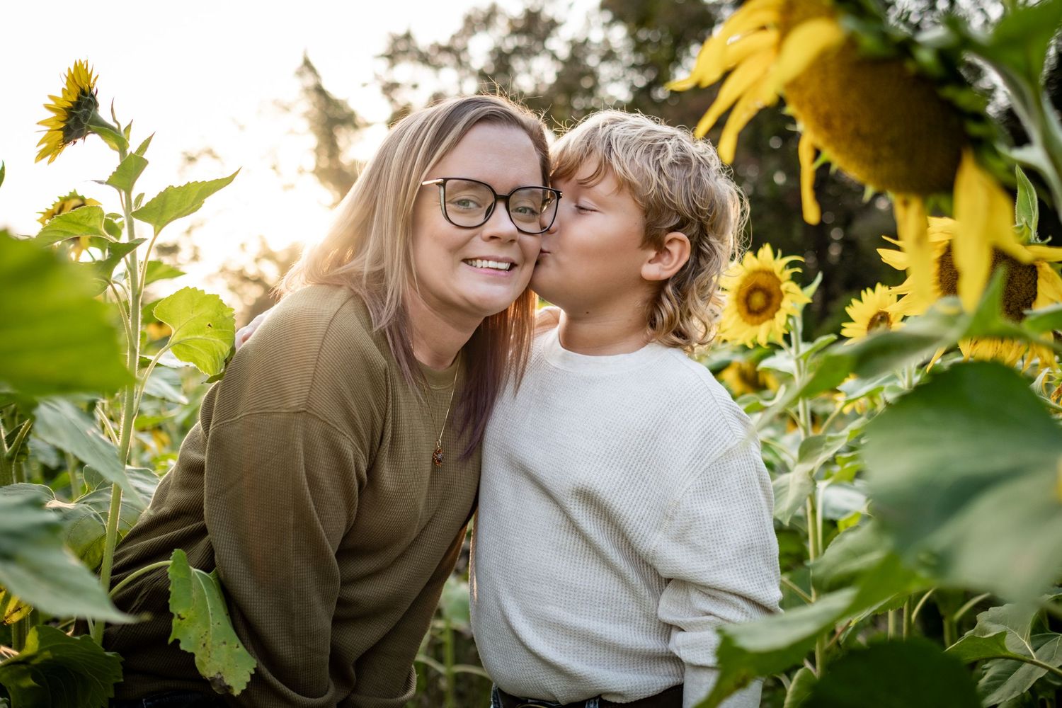 A child kisses a smiling woman wearing glasses among sunflowers in golden hour light.