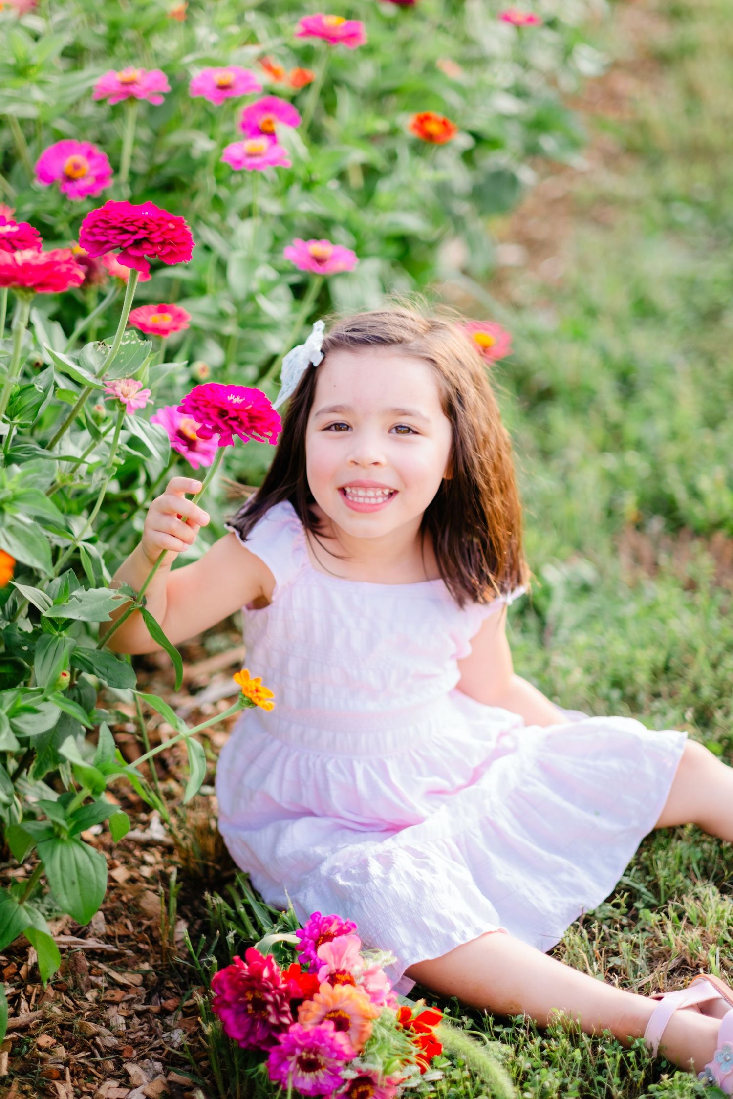 Young girl in pink dress sitting among colorful zinnia flowers in a garden, smiling and holding a pink bloom.