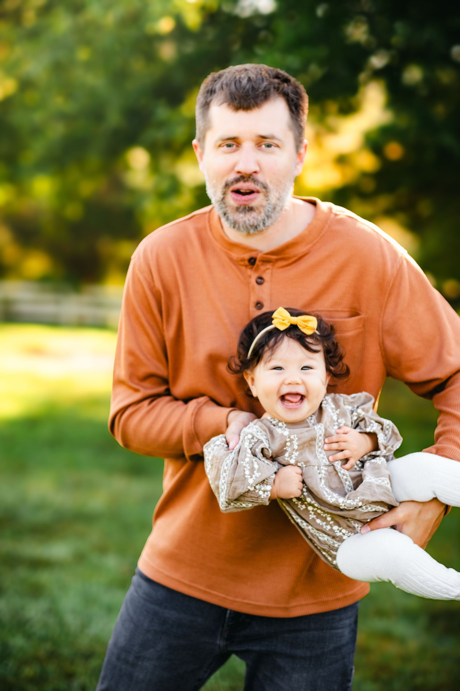 Man in rust orange shirt holding laughing toddler girl with yellow bow outdoors in green park setting.