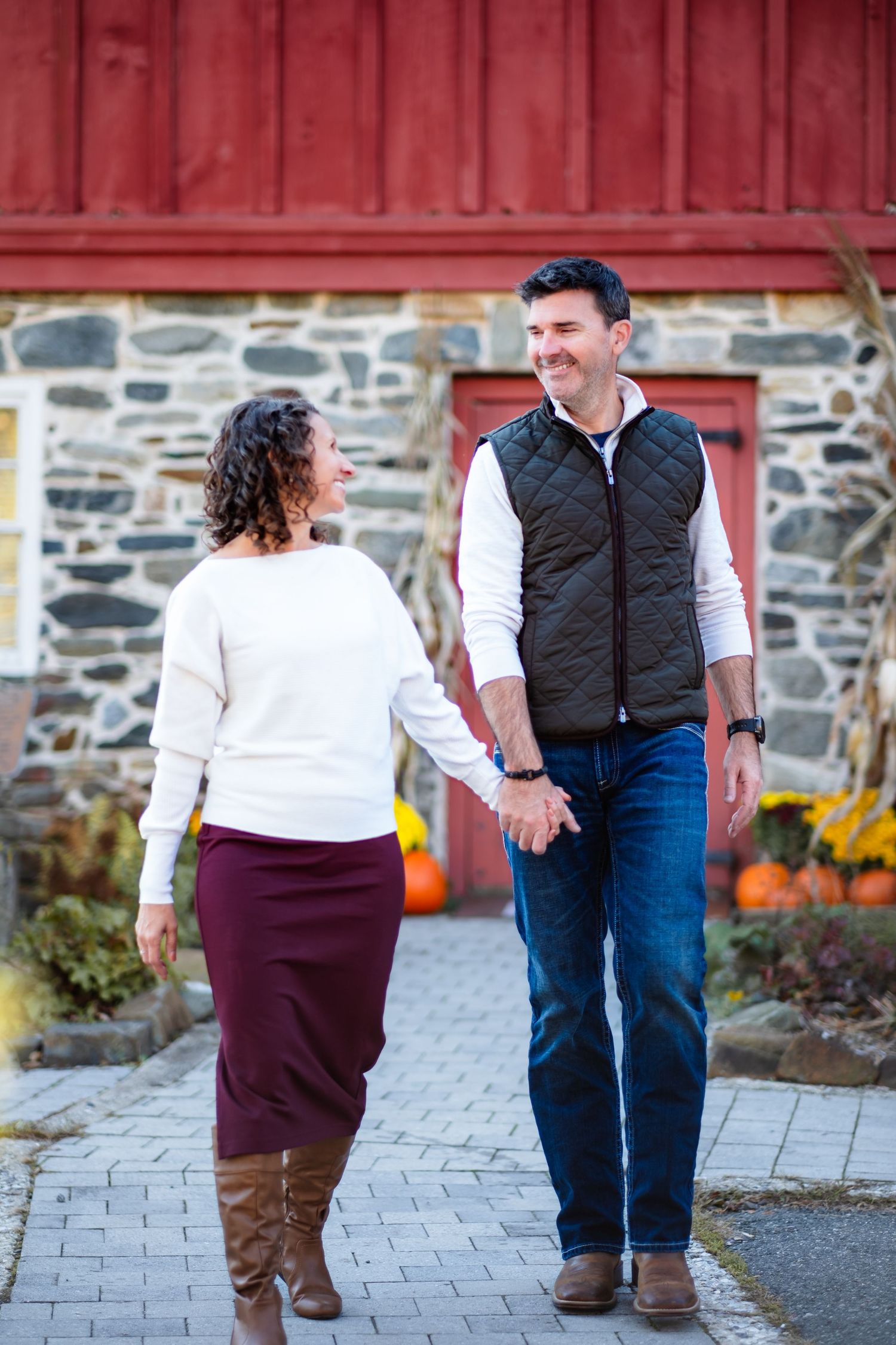 A couple holds hands and smiles at each other while walking outside a rustic stone barn in autumn.