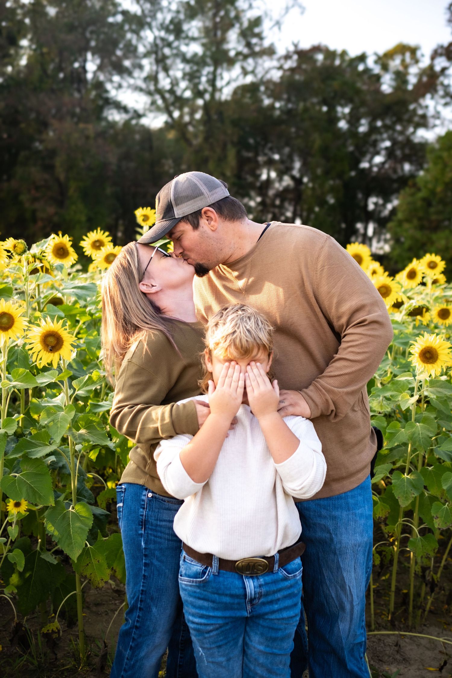 Family couple kissing in sunflower field while young child covers eyes, outdoor portrait photography.