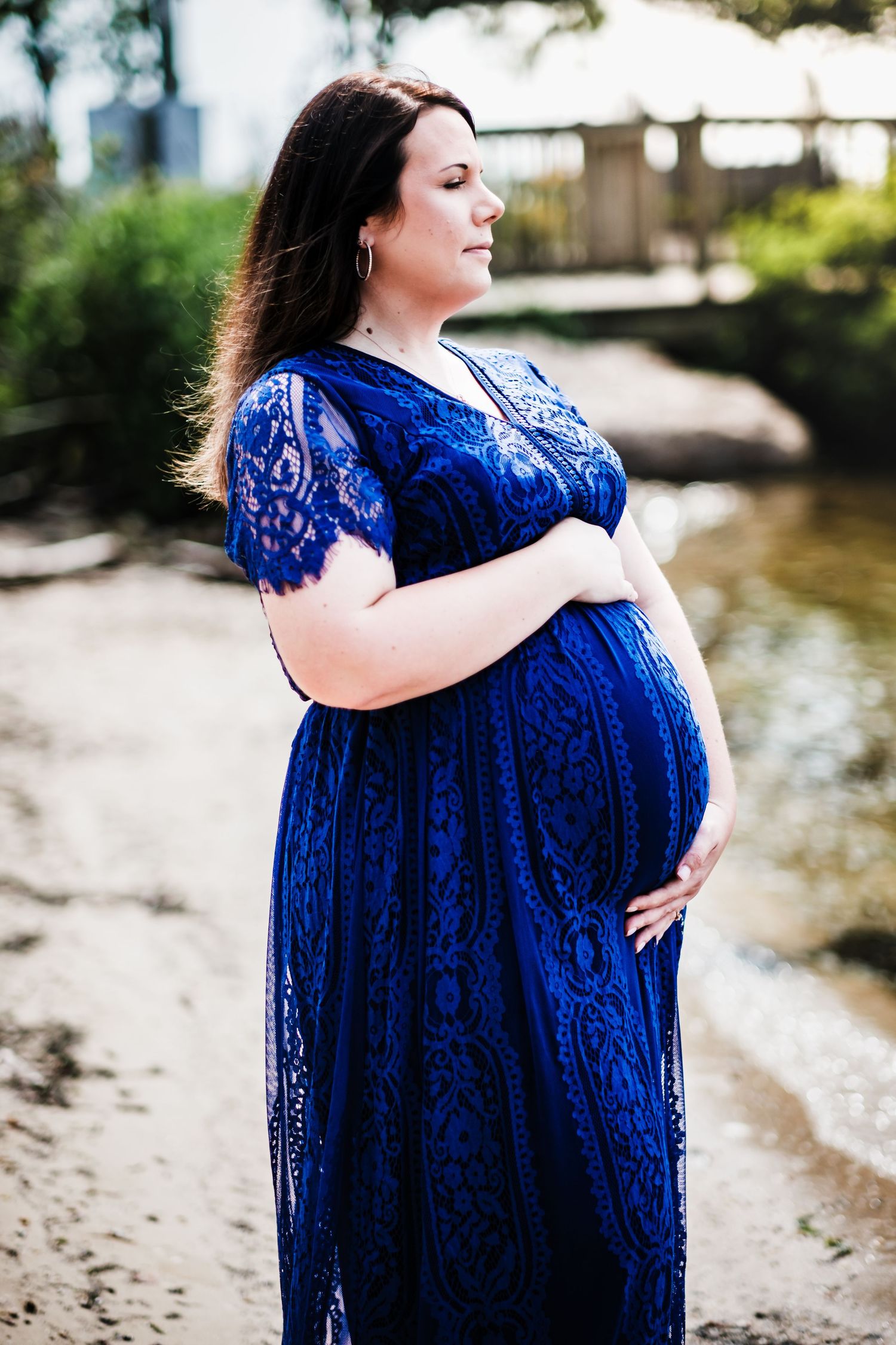 Pregnant woman in blue lace dress cradling baby bump near a serene riverside, maternity photography.