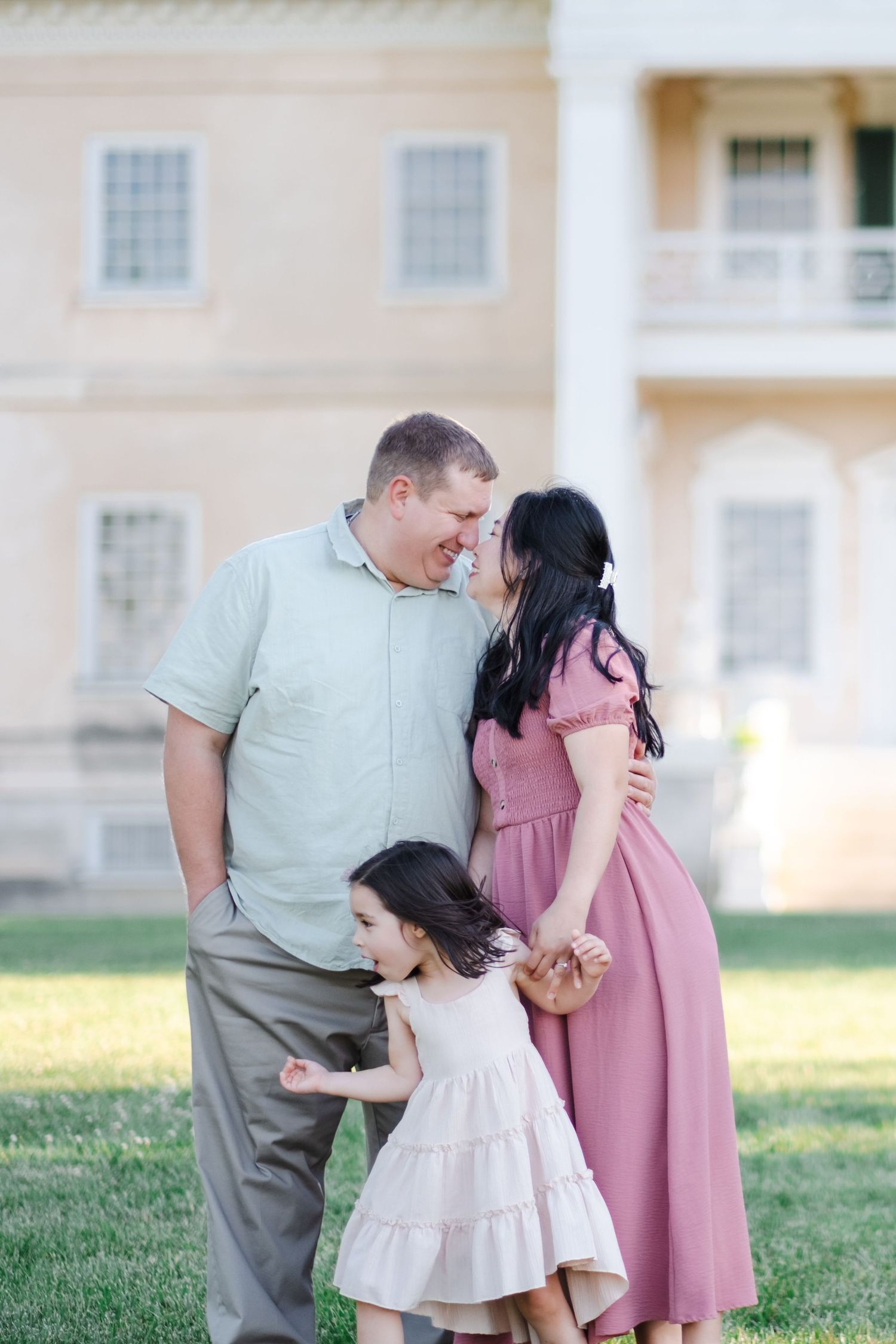 A happy family of three poses outdoors, parents smiling at each other while their young daughter plays between them.
