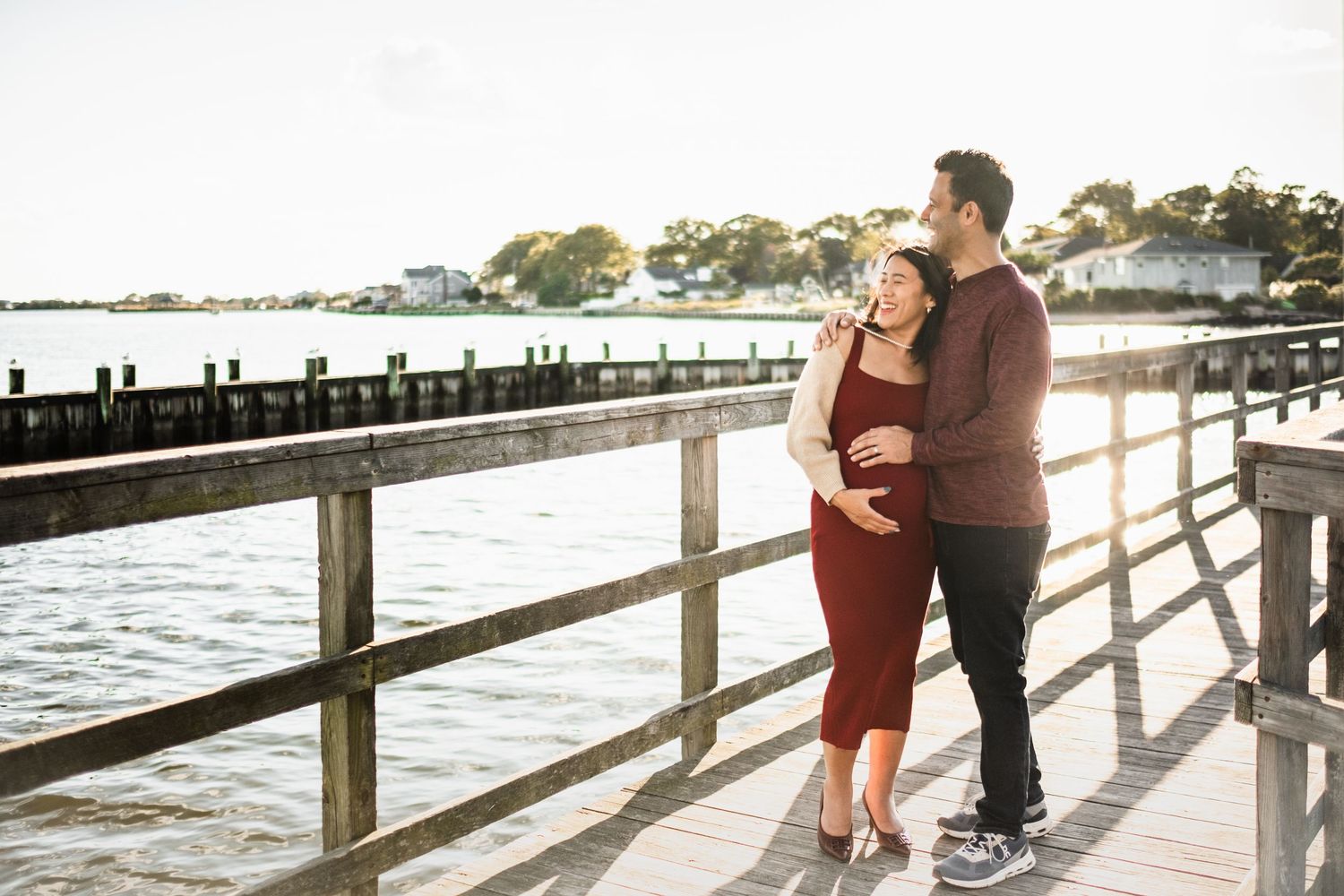 Pregnant couple embracing on a waterfront pier at golden hour, woman in red dress, man in burgundy sweater.