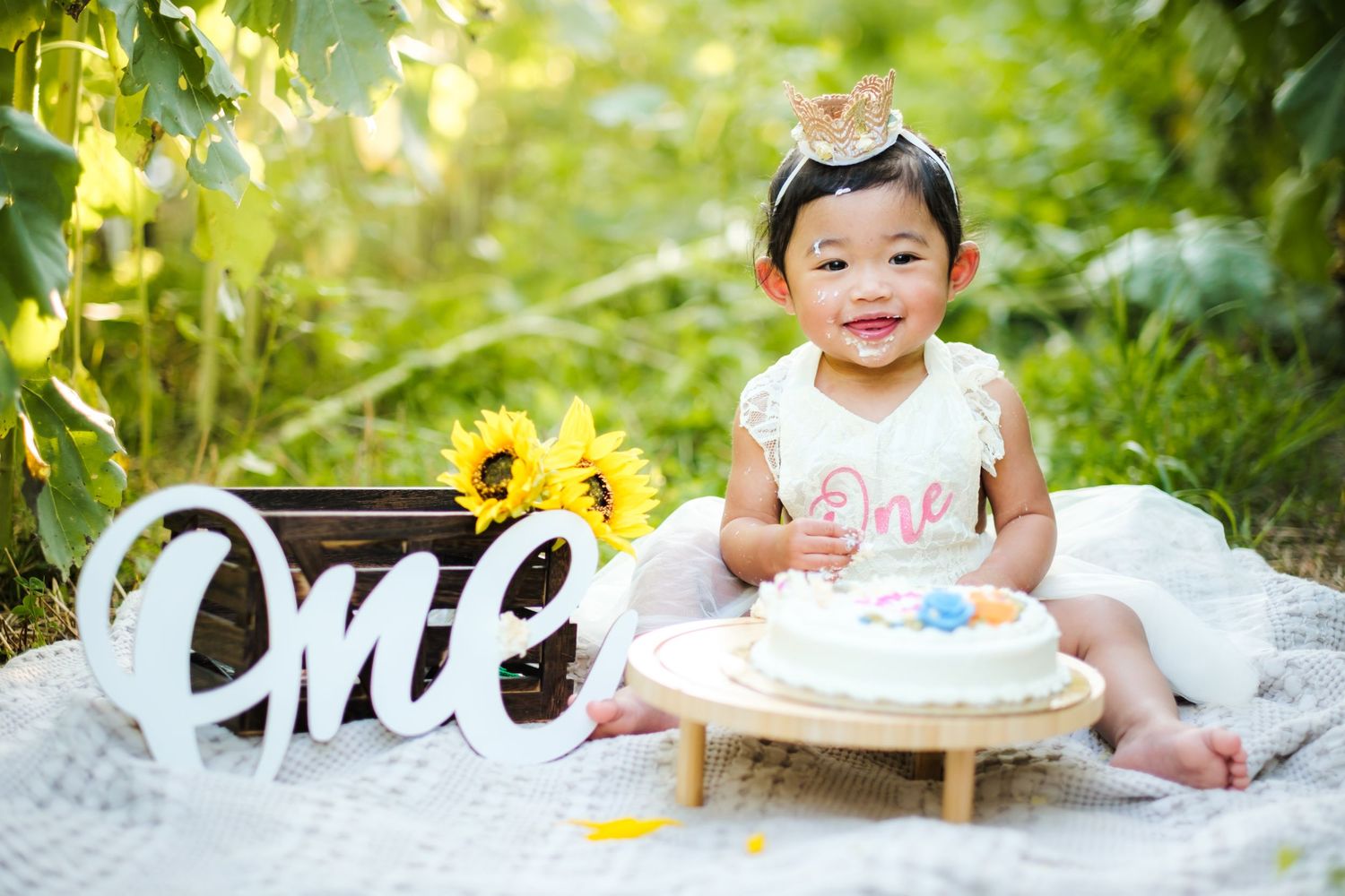 Baby girl in white dress celebrates first birthday outdoors with cake and sunflowers, wearing gold crown.