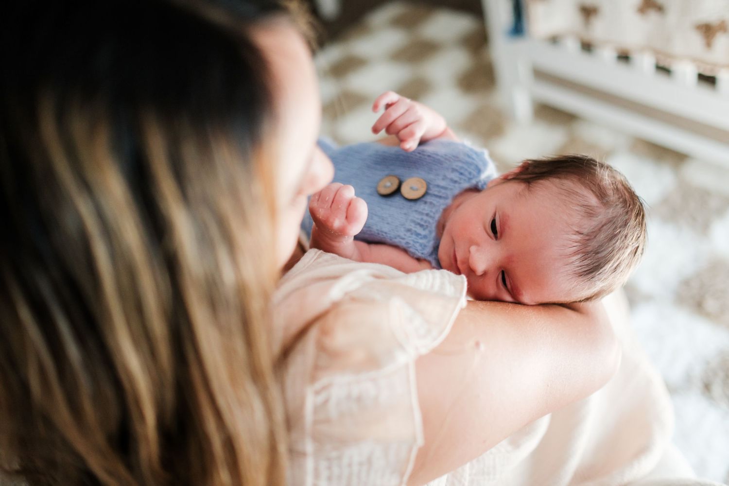 Mother tenderly holds sleeping newborn baby dressed in blue knit outfit during intimate lifestyle photography session.