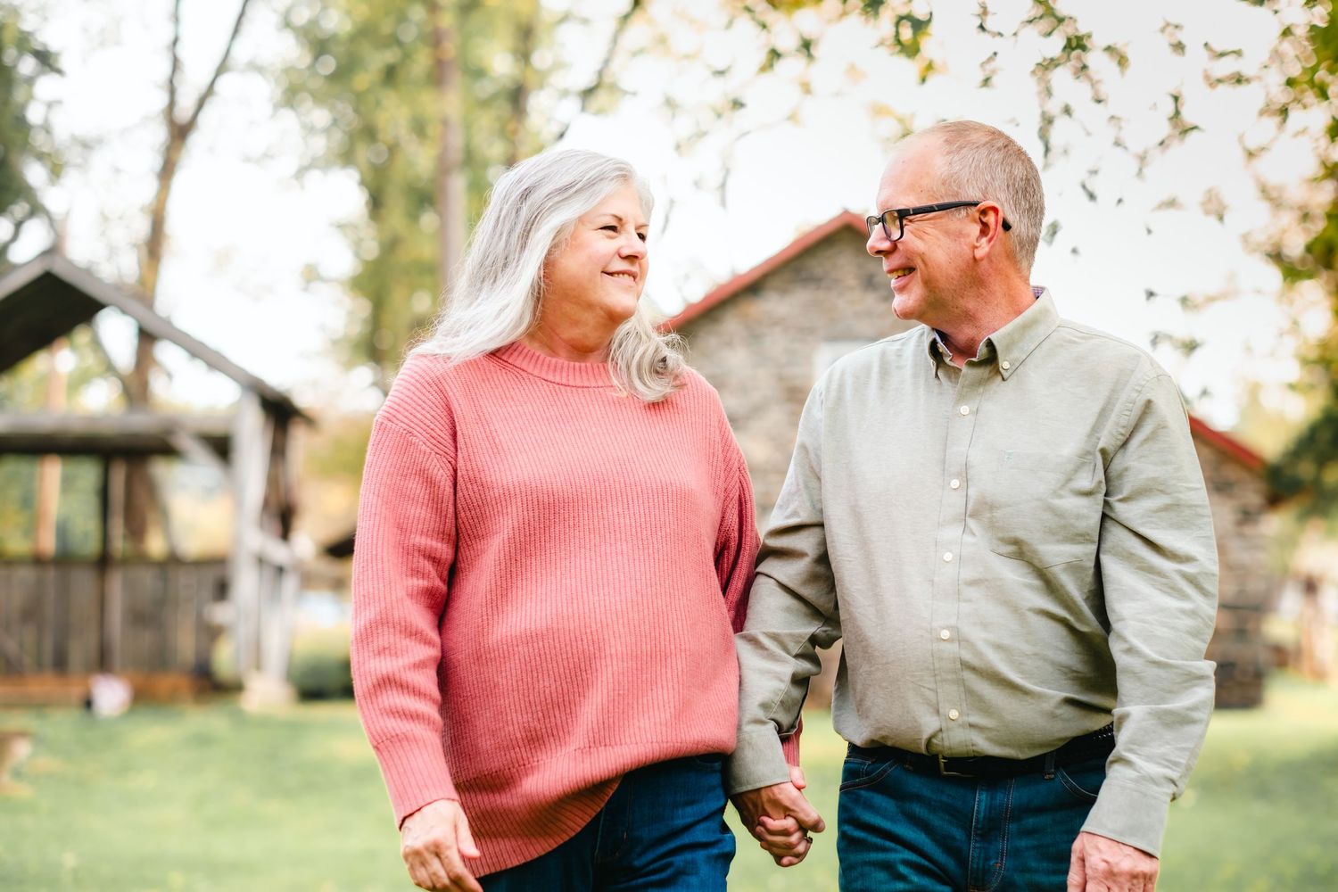 Happy senior couple holding hands and smiling at each other while walking outdoors in a lush green backyard.