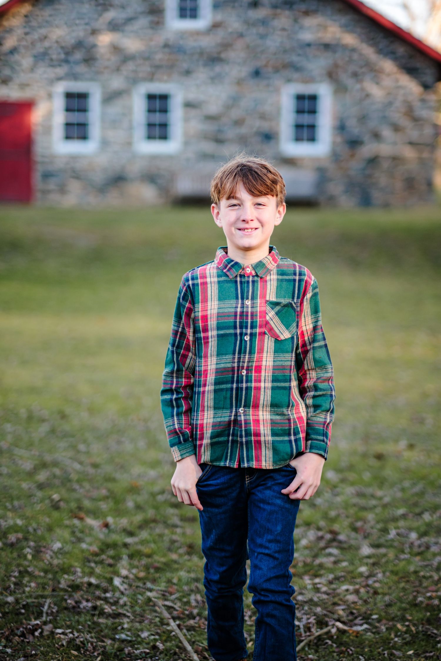 Smiling boy in green plaid shirt and jeans standing outdoors in front of a historic stone building.