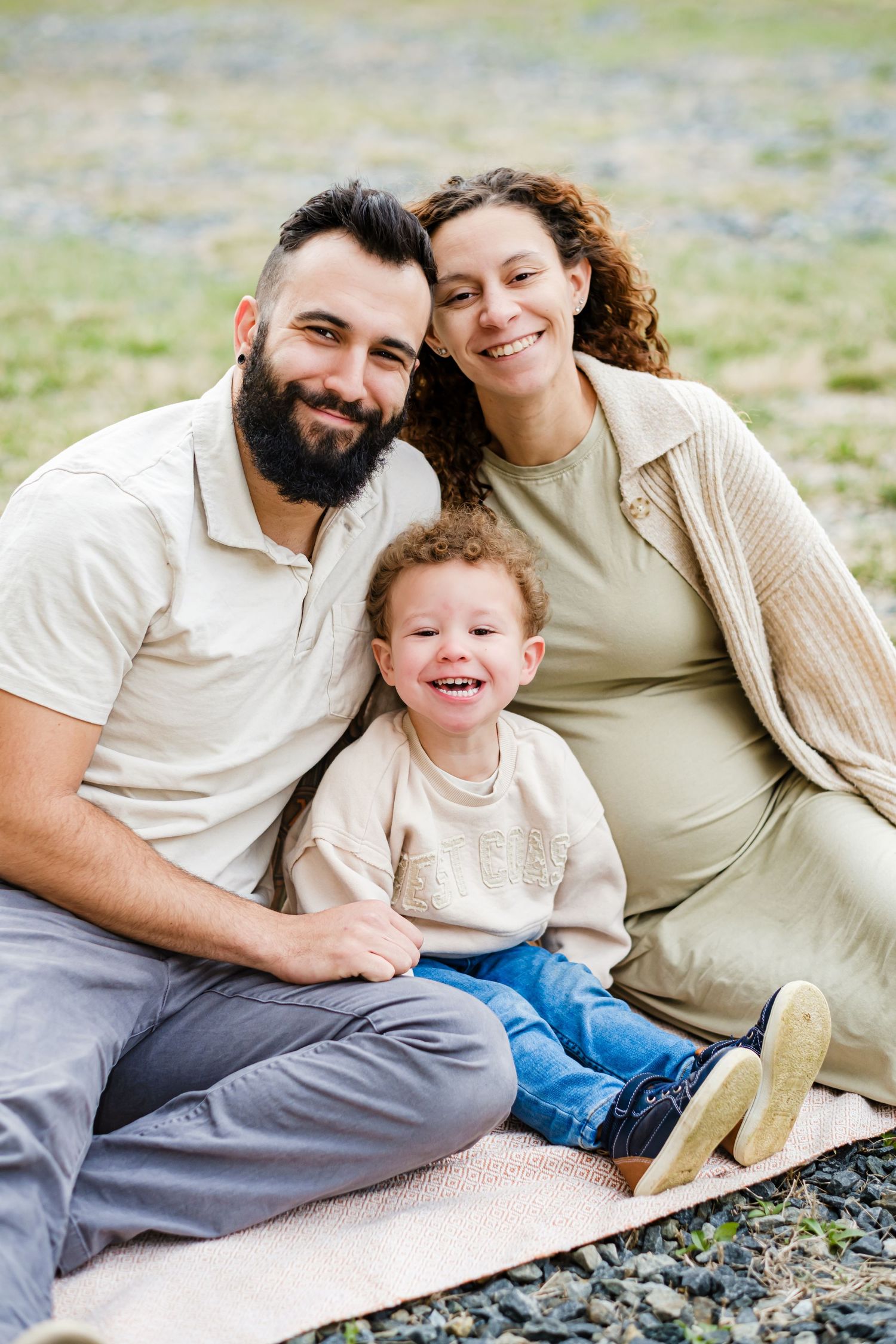 Happy family of three sitting on a blanket outdoors, with bearded dad, curly-haired mom, and laughing toddler.