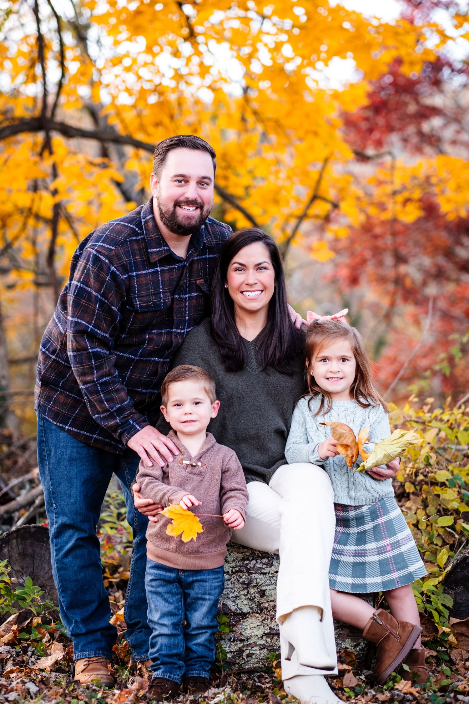 Family of four posing outdoors among vibrant autumn foliage, smiling warmly in coordinated fall outfits.
