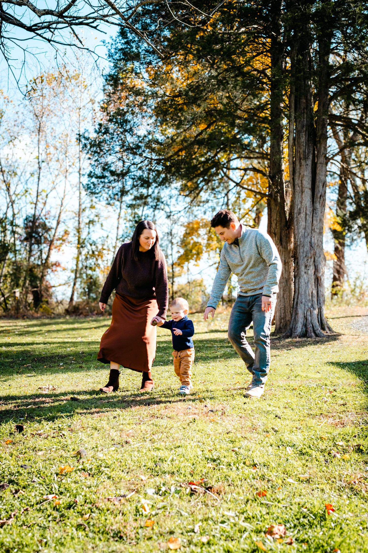 A young family with a toddler enjoys a sunny autumn day in a lush green park surrounded by colorful fall trees.