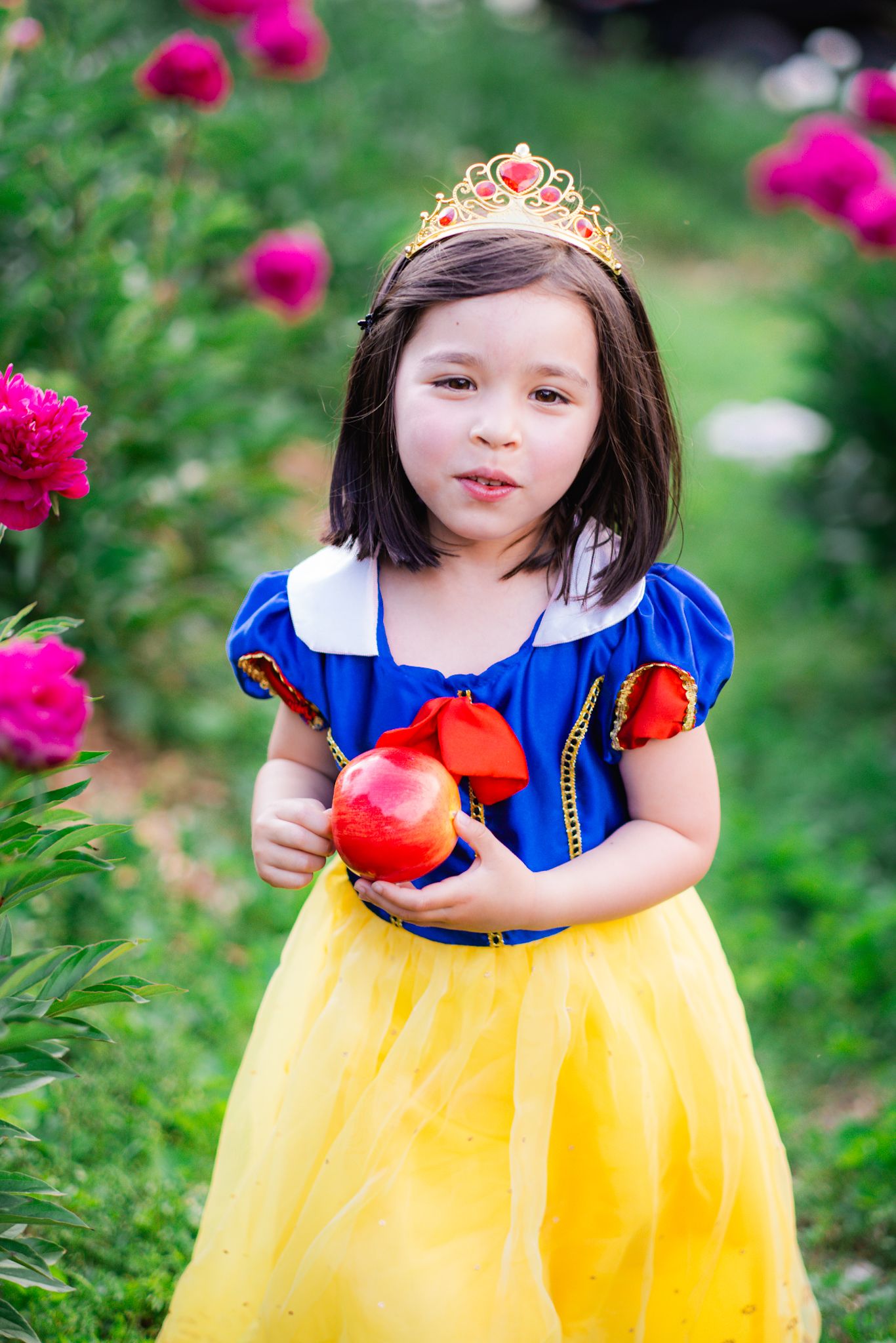 A little girl dressed as Snow White holds a red apple in a garden with pink flowers.