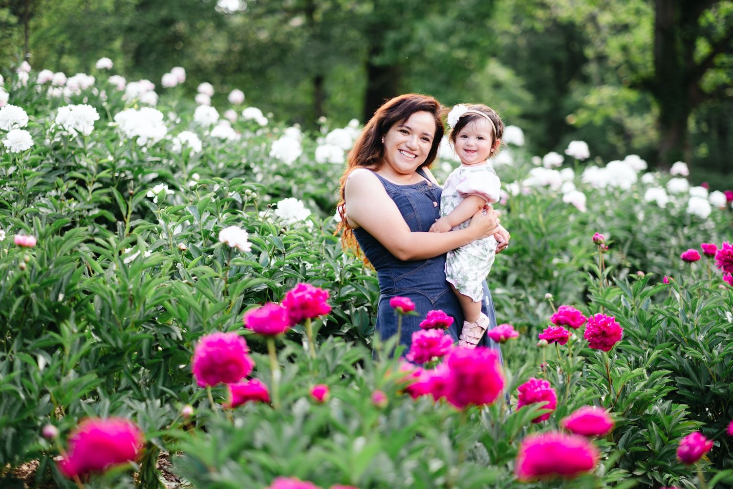 A woman holding a toddler smiles in a vibrant garden filled with pink and white peony blooms.