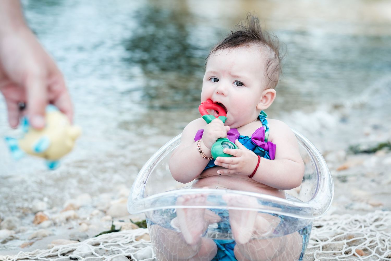 Adorable baby sitting in a clear bowl on a rocky beach, chewing a colorful toy while an adult hand offers a snack.