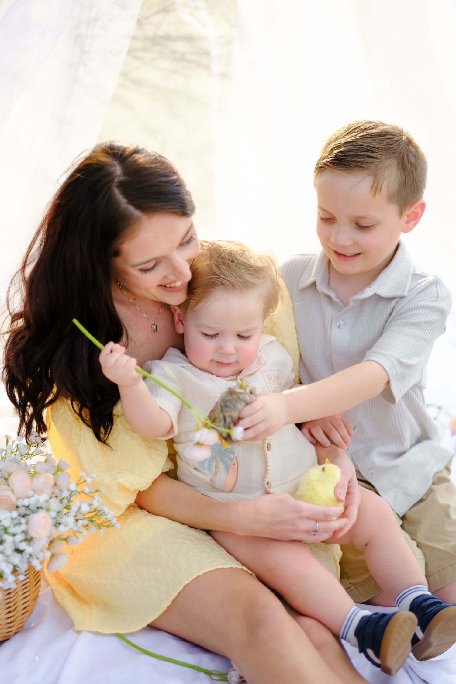 Happy mother in yellow dress sits with two young sons examining Easter chick on white blanket outdoors.