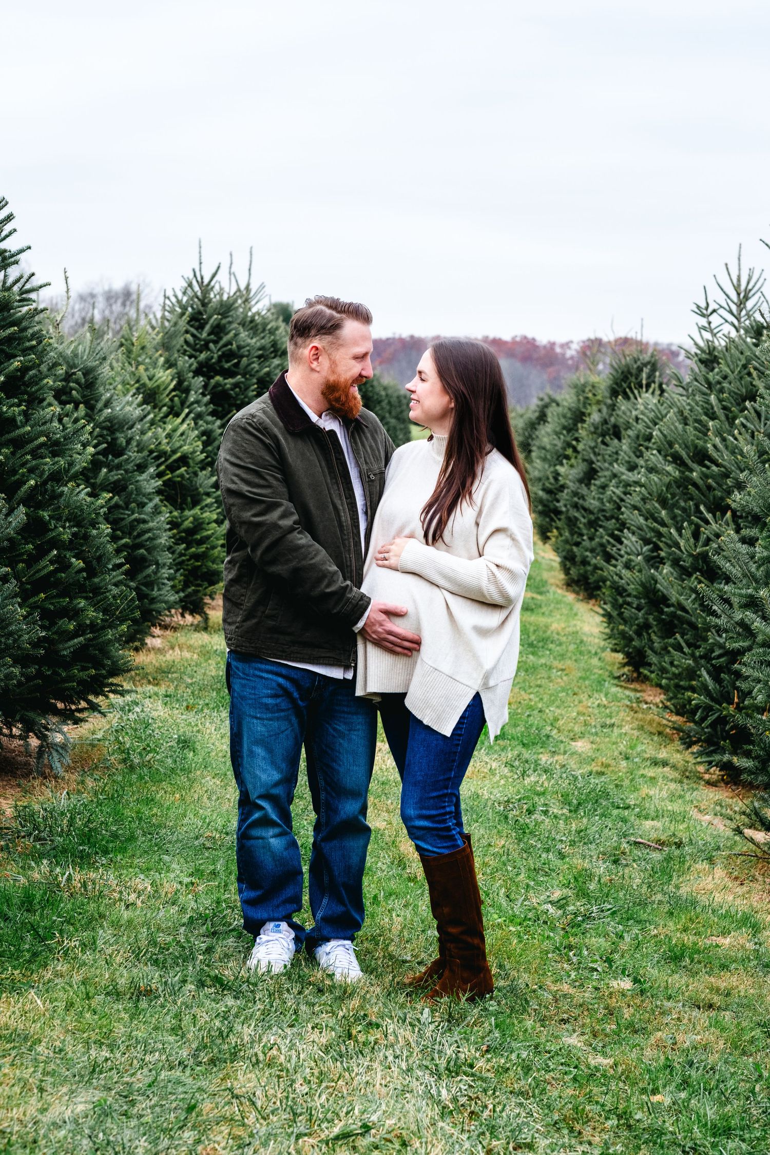 Couple sharing a tender moment at a Christmas tree farm, woman visibly pregnant, surrounded by rows of evergreen trees.