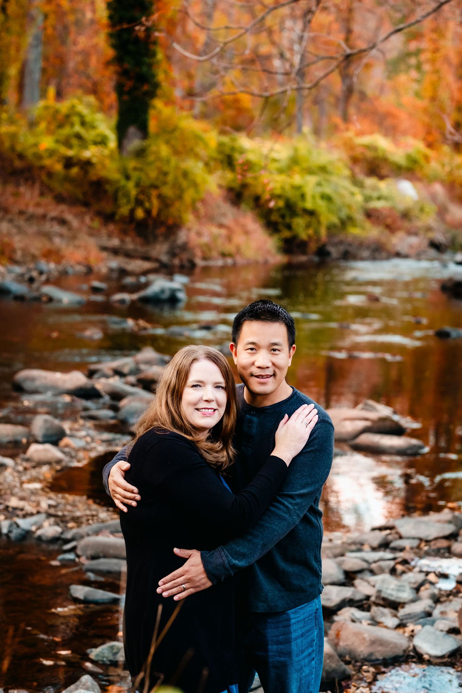 Couple embracing by a rocky stream surrounded by vibrant autumn foliage in warm orange and red tones.