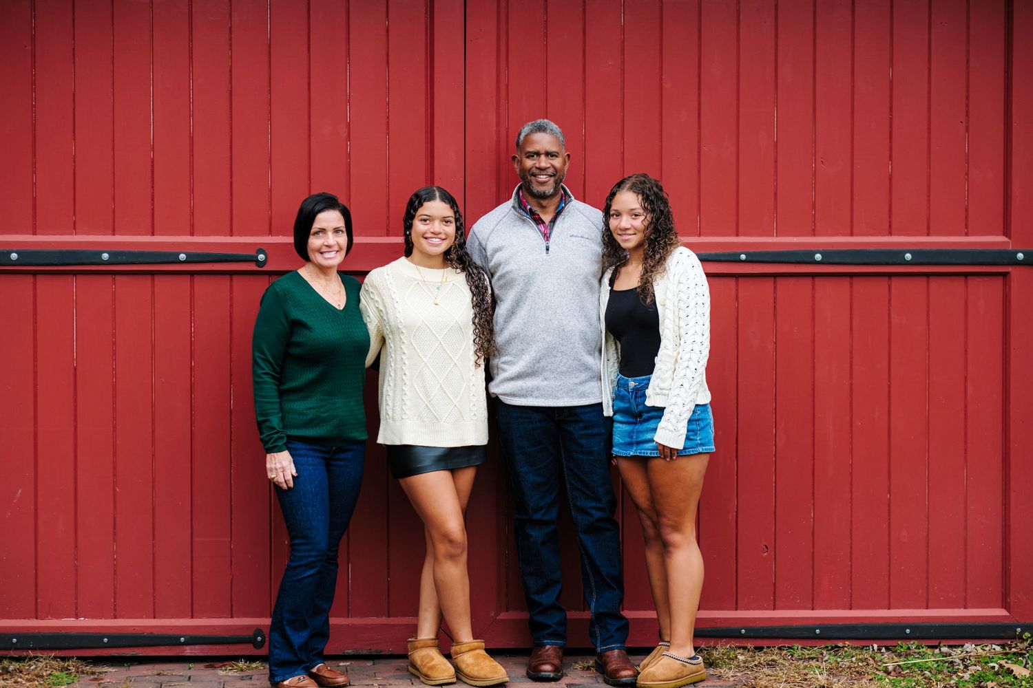 Family of four smiling together in front of a red barn wall outdoors.