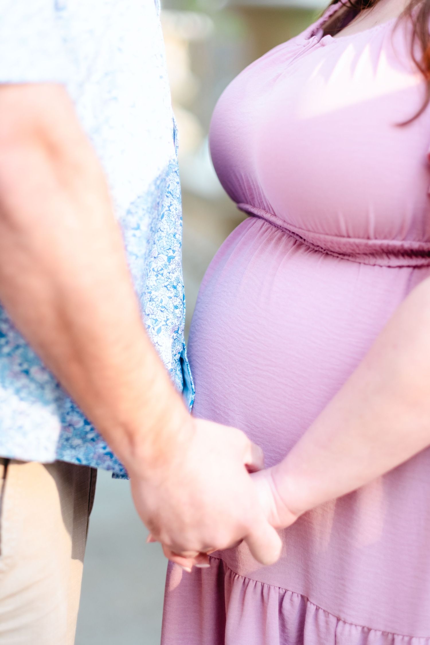 Pregnant woman in purple dress holding hands with partner, showcasing baby bump during maternity photoshoot outdoors.