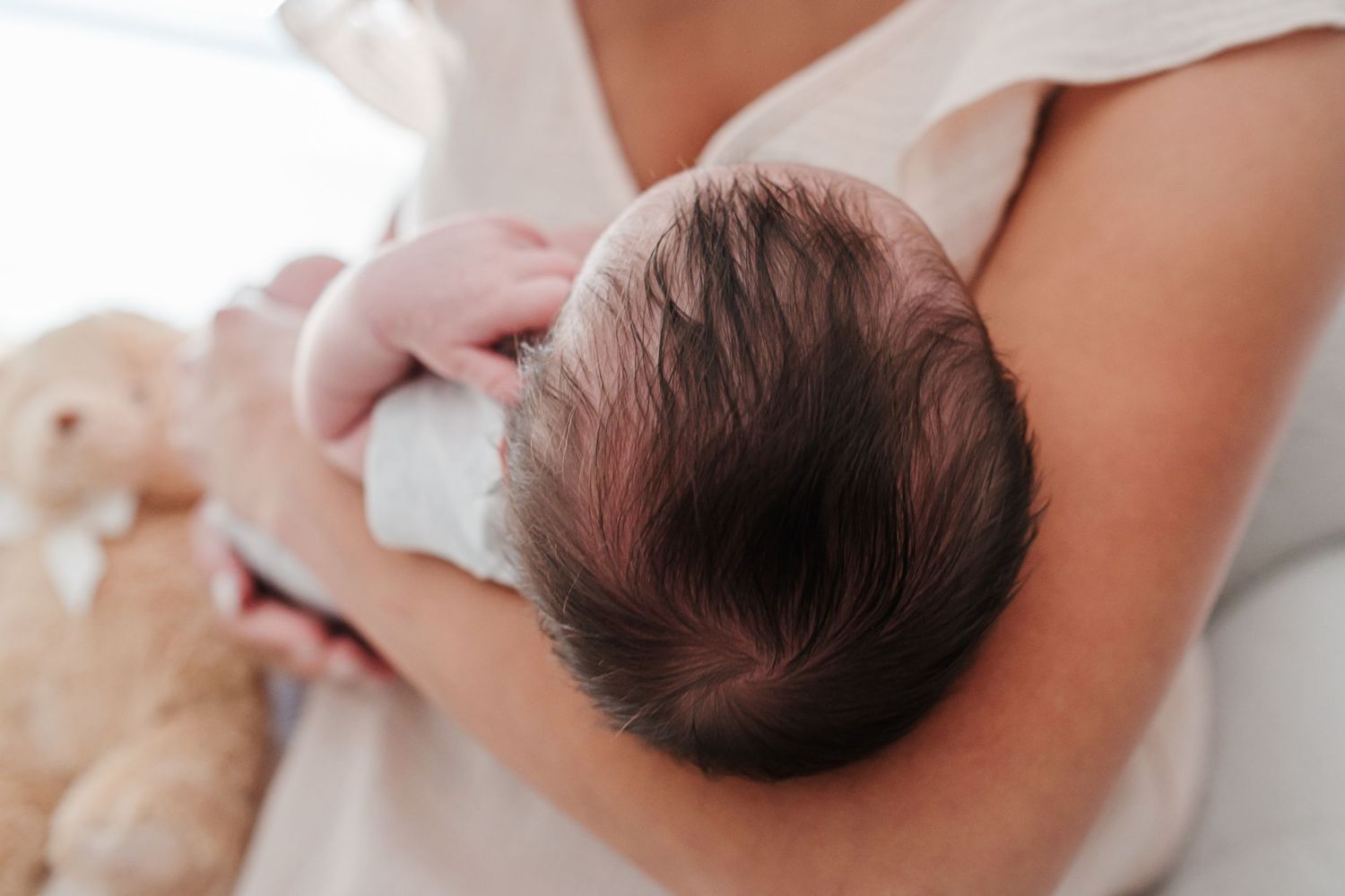 Mother breastfeeding her newborn baby with dark hair, held close to her chest near a soft teddy bear.