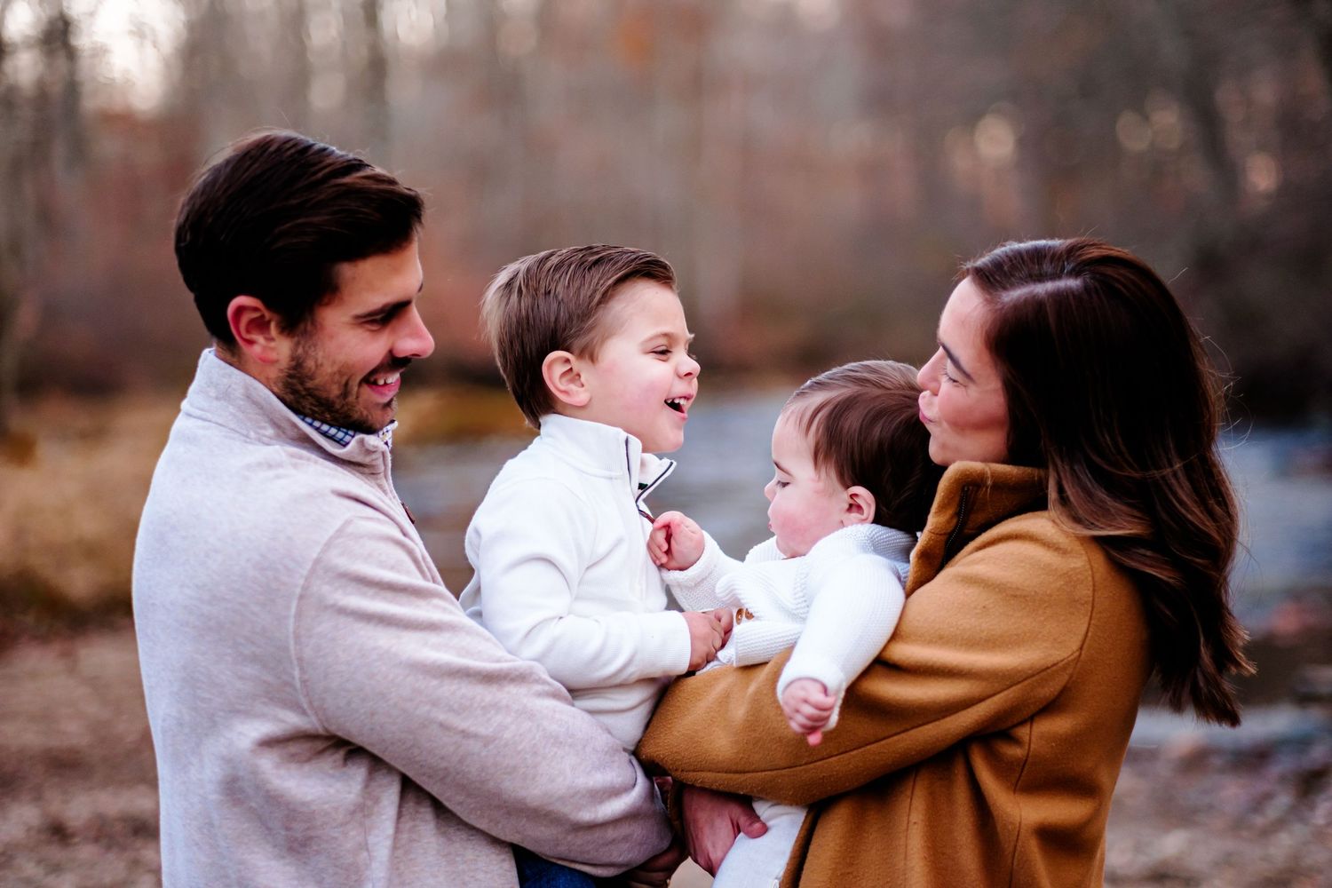 A happy family of four poses outdoors in autumn, parents holding two young children, smiling warmly in neutral tones.