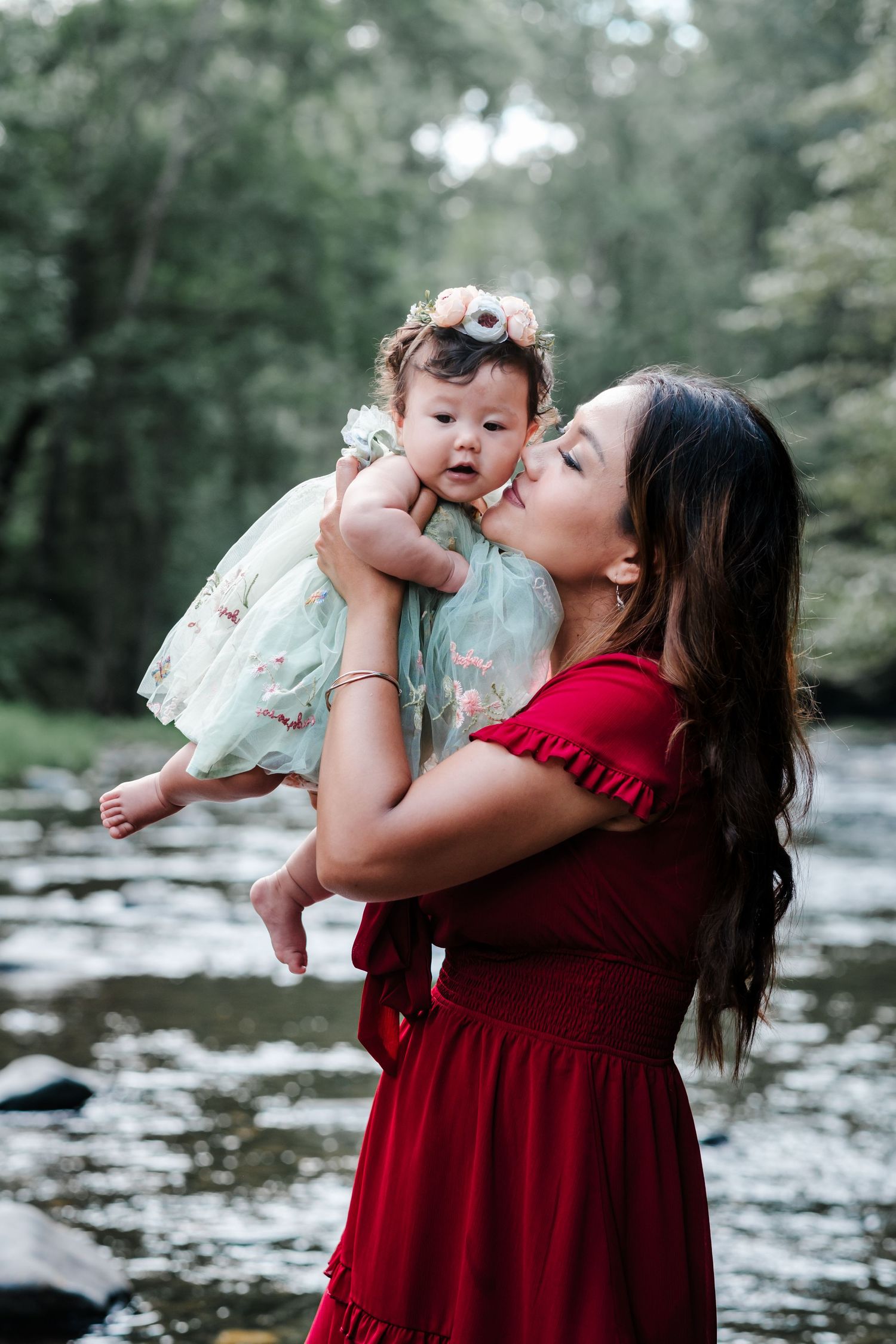 A mother in a red dress kisses her baby girl in a floral dress by a serene forest stream.