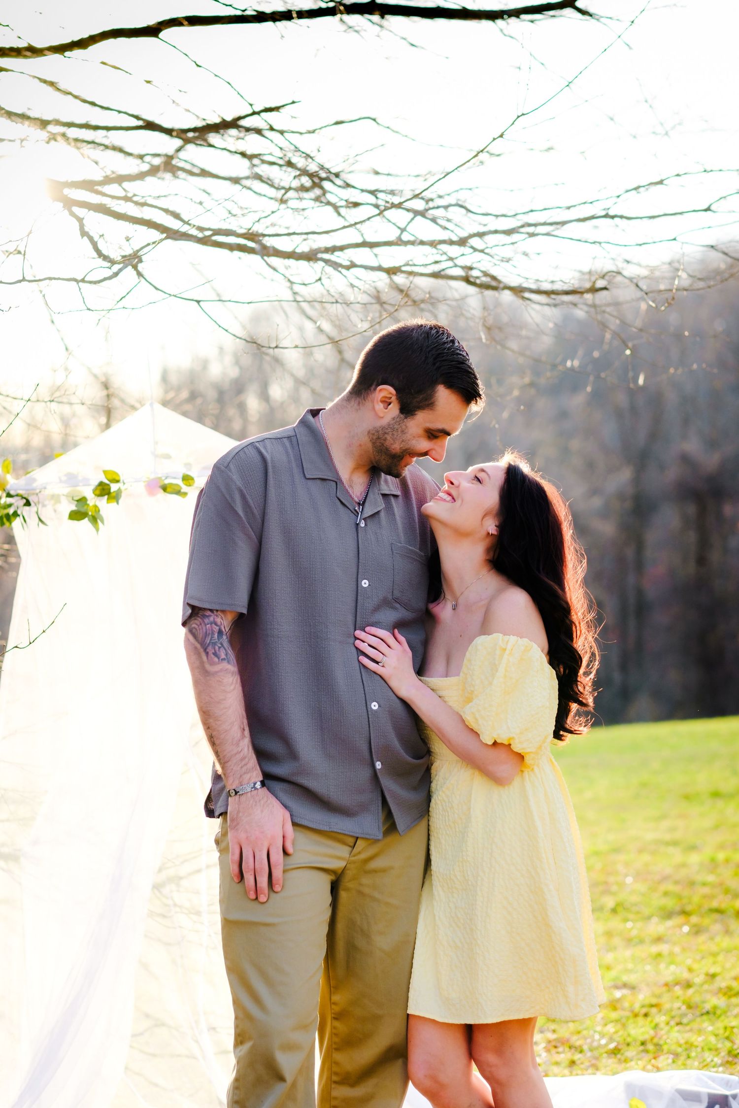 Romantic couple embraces and laughs together outdoors under bare tree branches in warm spring sunlight.