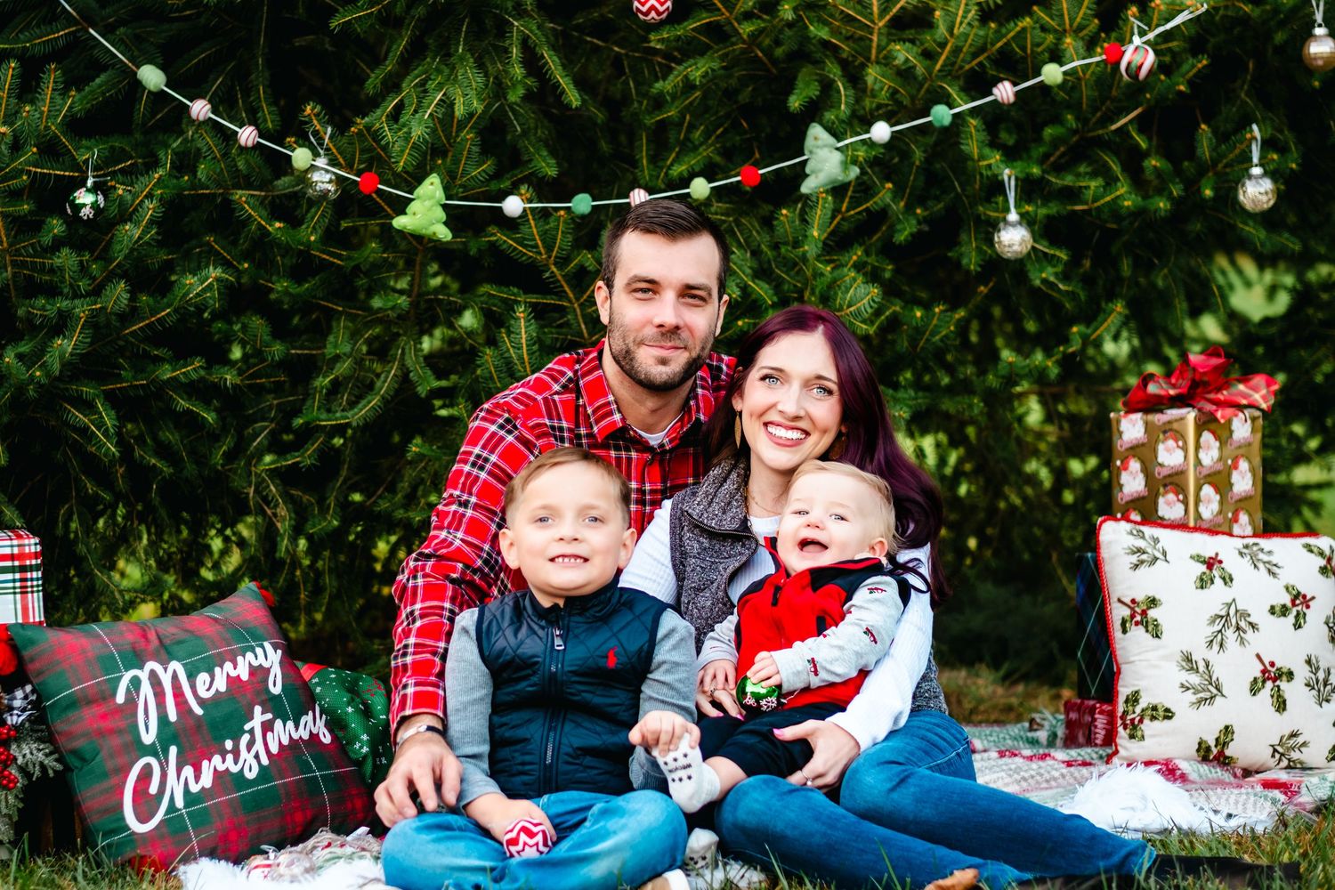 Family of four poses for a festive Christmas photo outdoors with gifts and holiday decorations.
