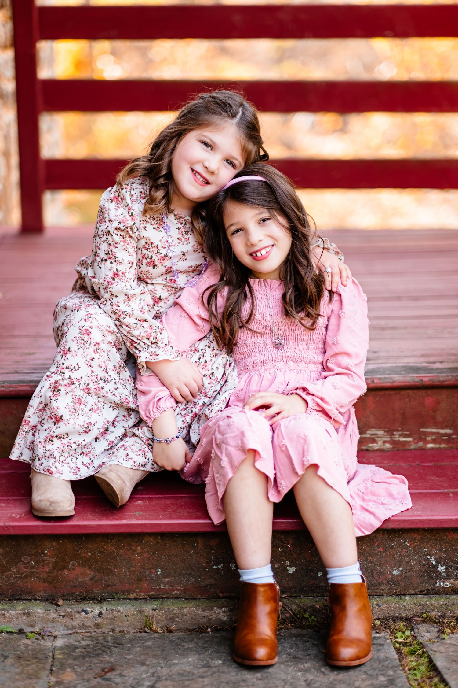 Two young girls in pink dresses and brown boots sit together on red steps, smiling warmly in an autumn setting.