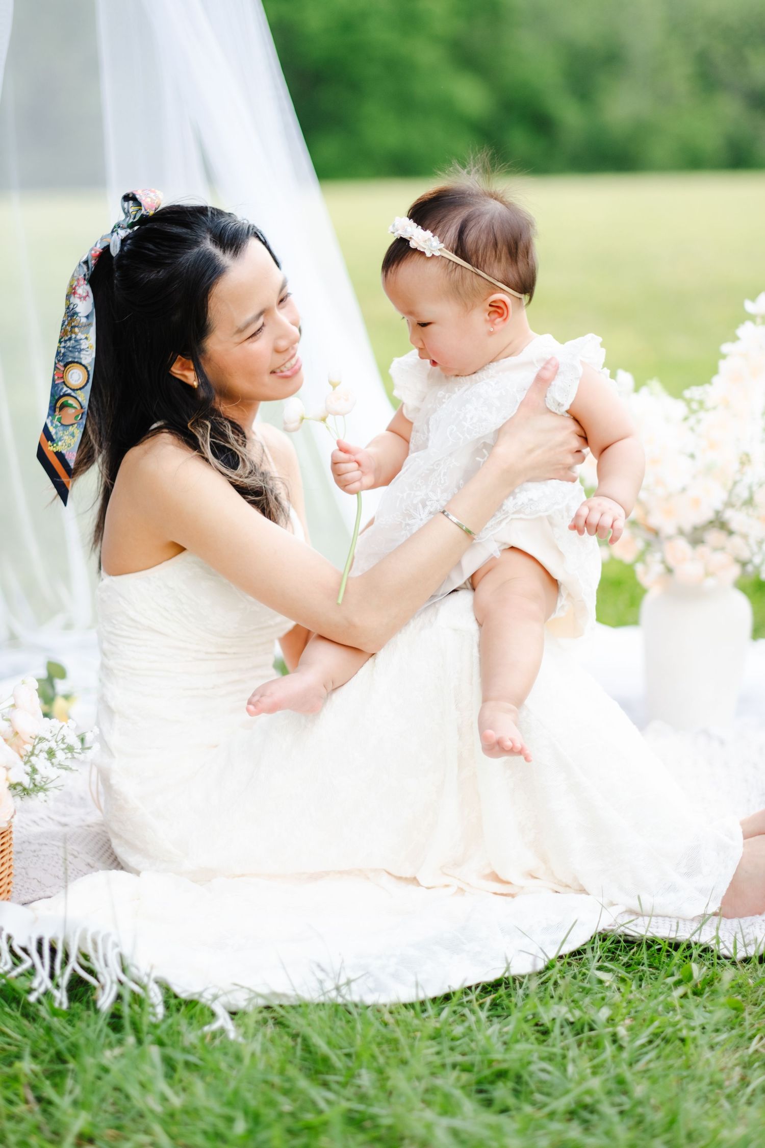 A smiling Asian mother in white dress holds her baby girl in white outfit on a blanket outdoors.