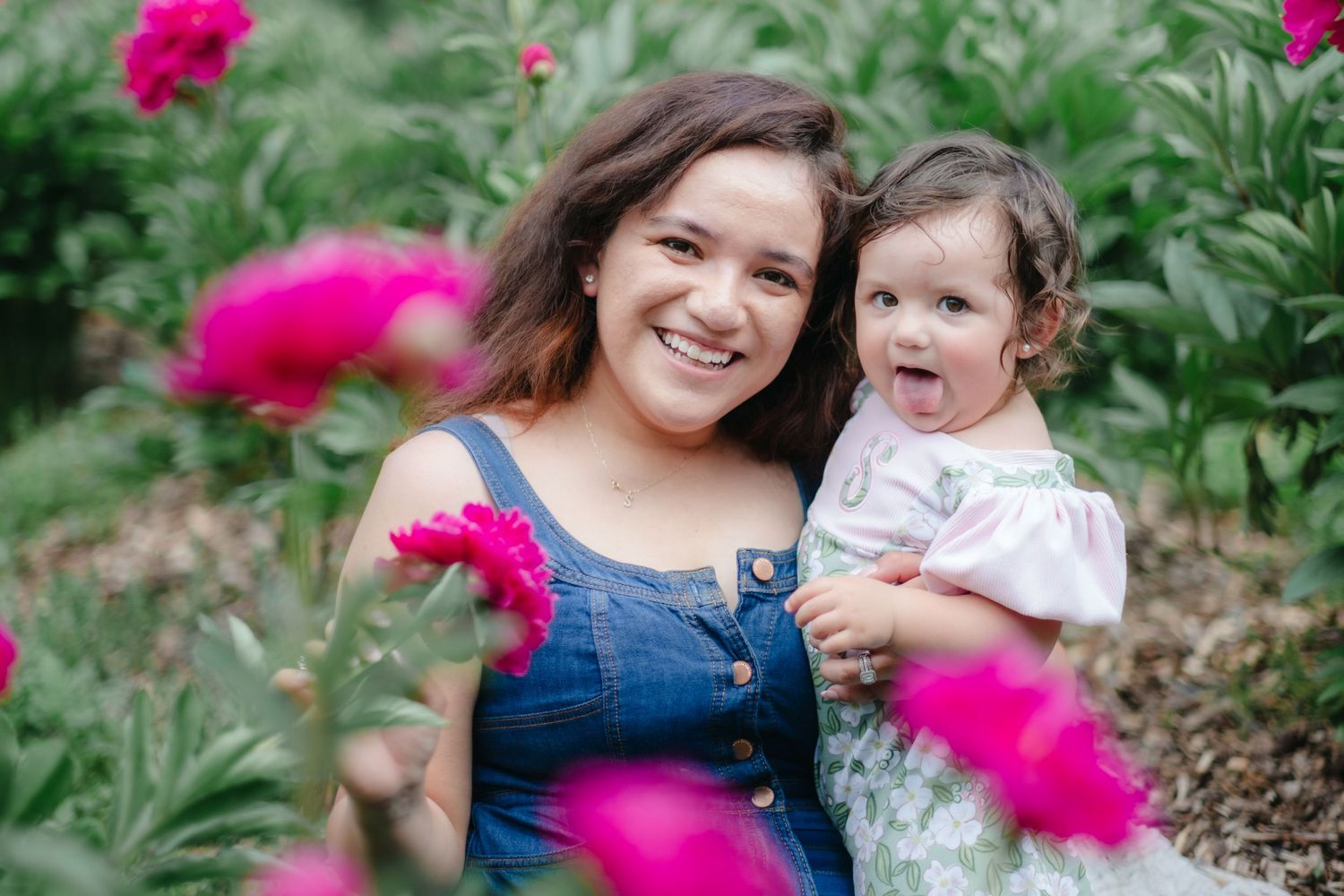 A young woman in a denim dress smiles holding a toddler girl among vibrant pink peonies in a lush garden.