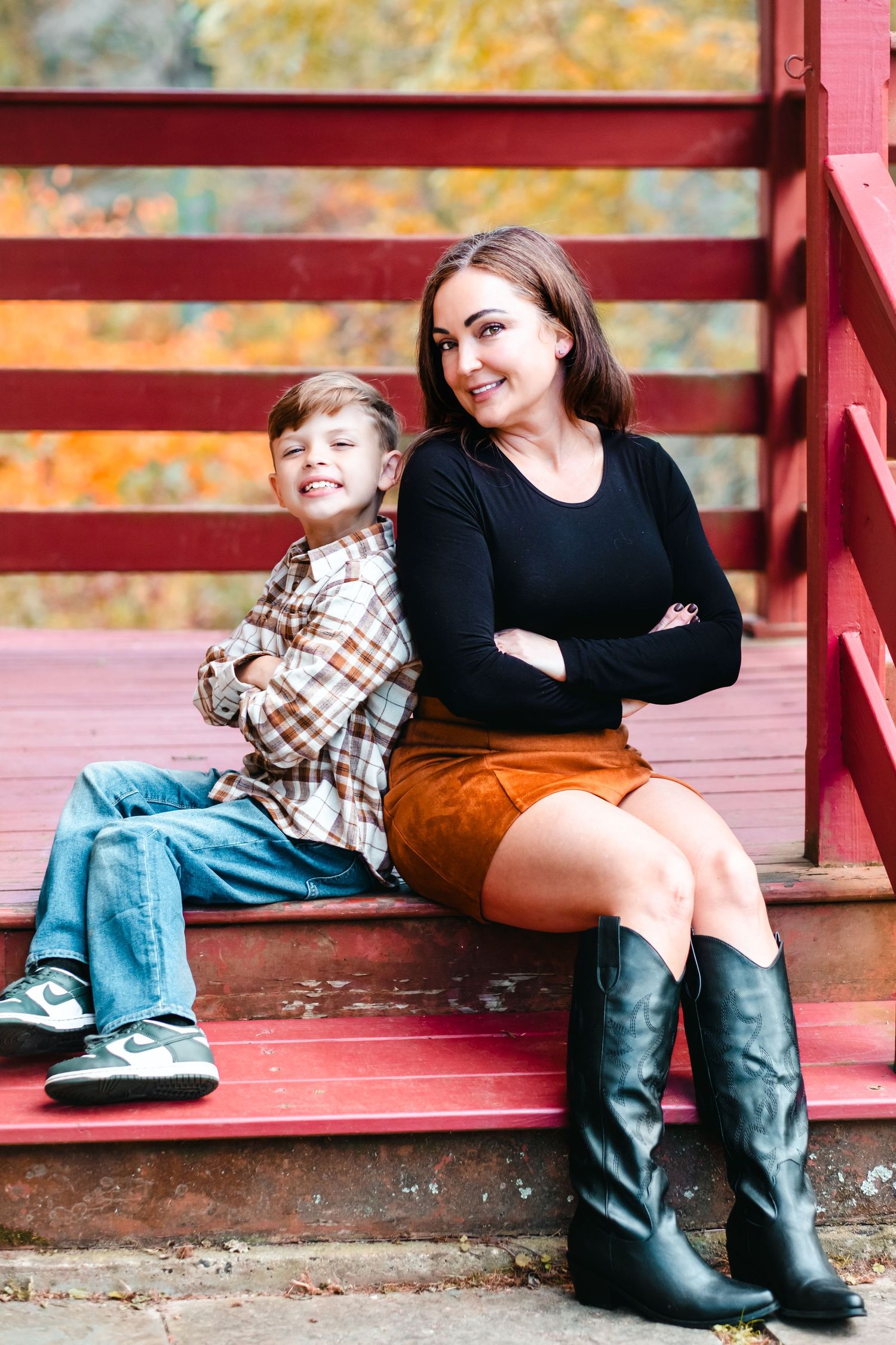 A young woman in a black top, suede skirt, and boots sits with a boy on red steps, both smiling with arms crossed.