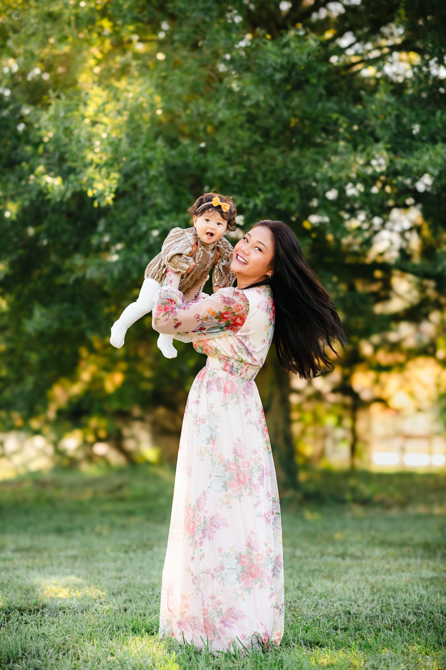 A smiling mother in a floral maxi dress lifts her baby girl in a sunny green park.