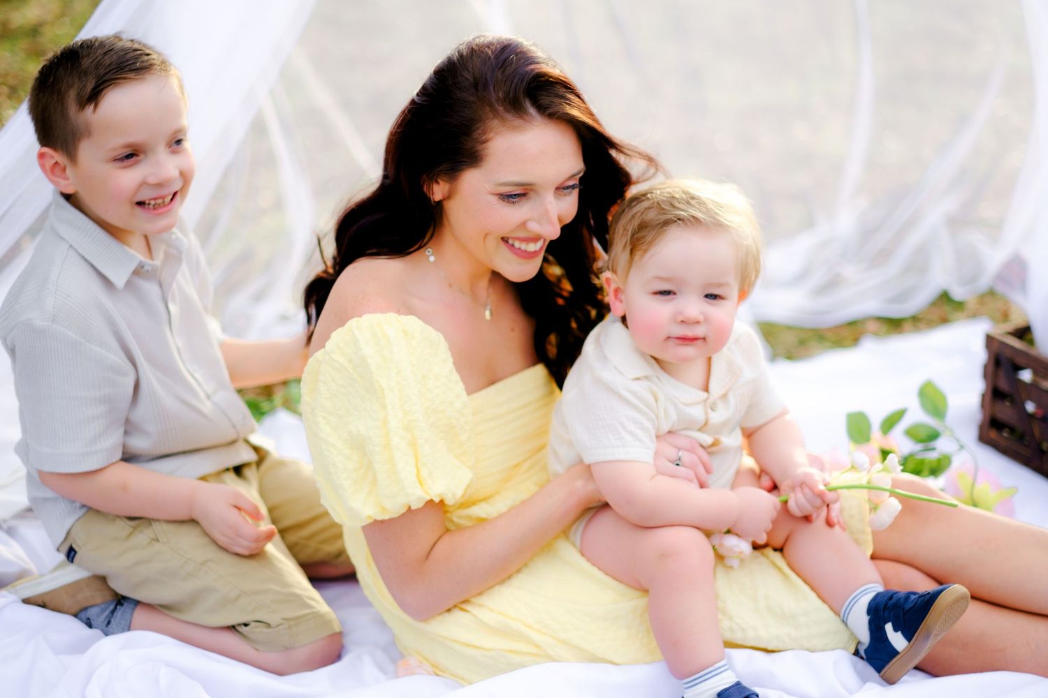 A smiling mother in a yellow dress sits outdoors with her two young sons, enjoying a bright sunny day together.