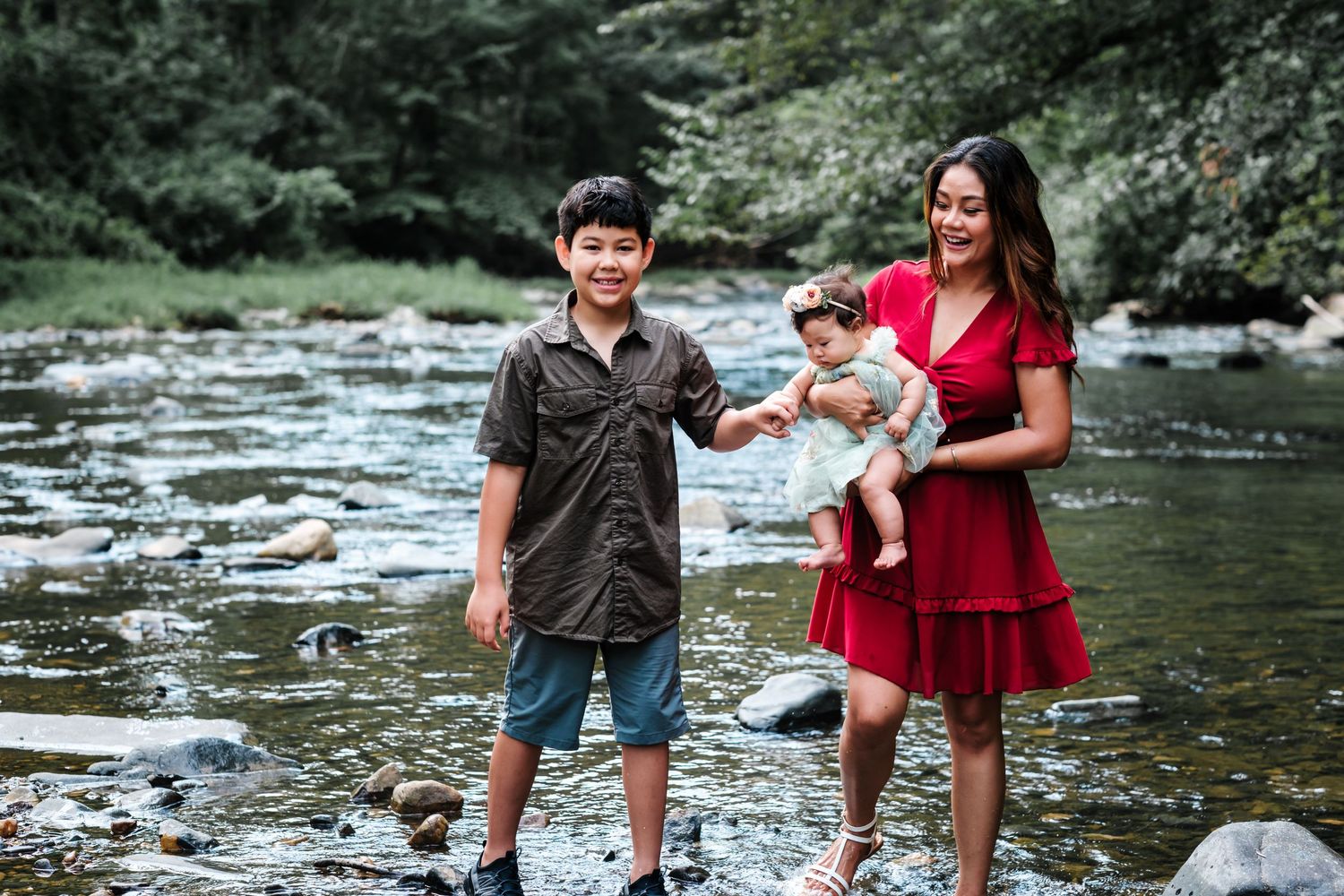 A mother in a red dress holds a baby while standing with her young son on rocky riverbank surrounded by lush greenery.