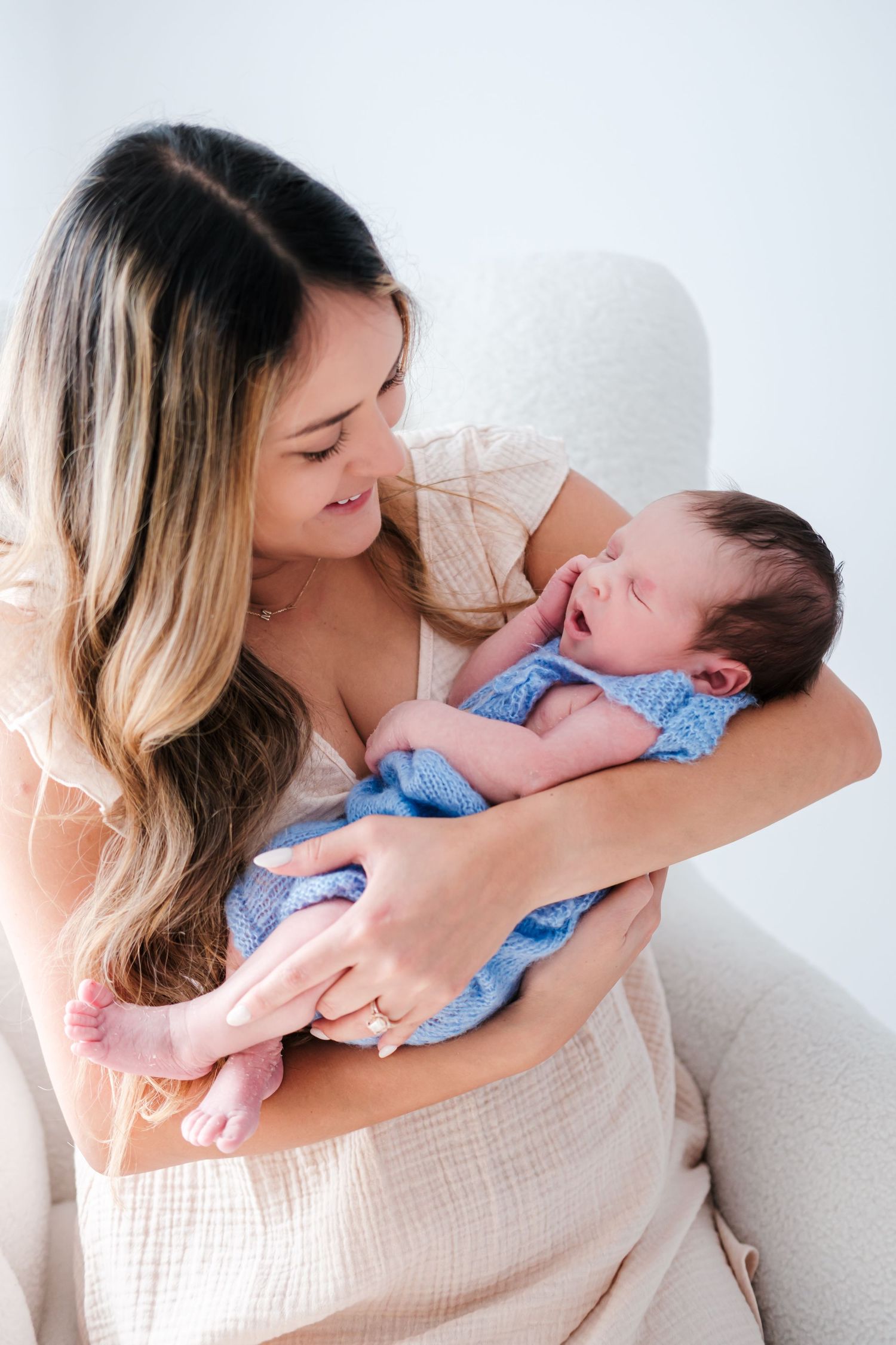 A mother tenderly holds her sleeping newborn baby wrapped in a blue knit blanket while sitting in a white chair.