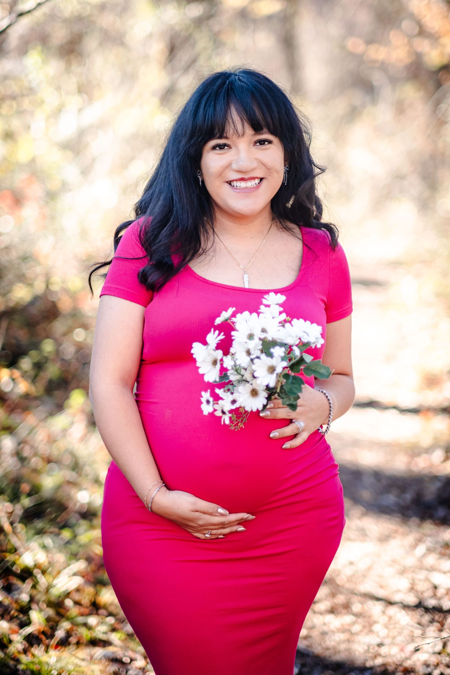 Smiling pregnant woman in pink dress holding white flowers outdoors in a sunlit forest maternity photo.