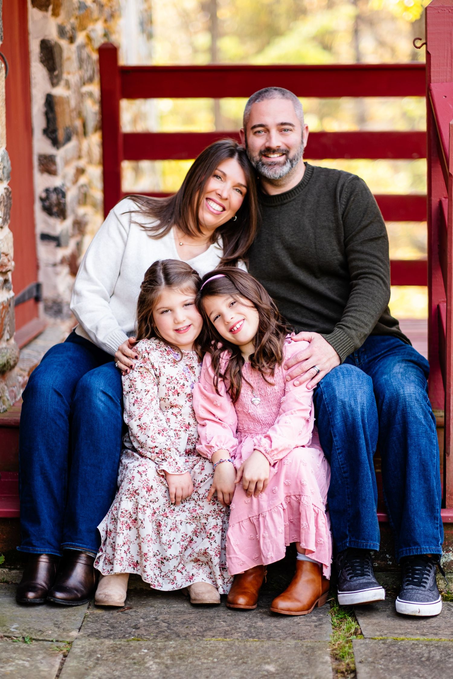 A smiling family of four poses on red steps, two parents and two young girls in floral dresses, during autumn.