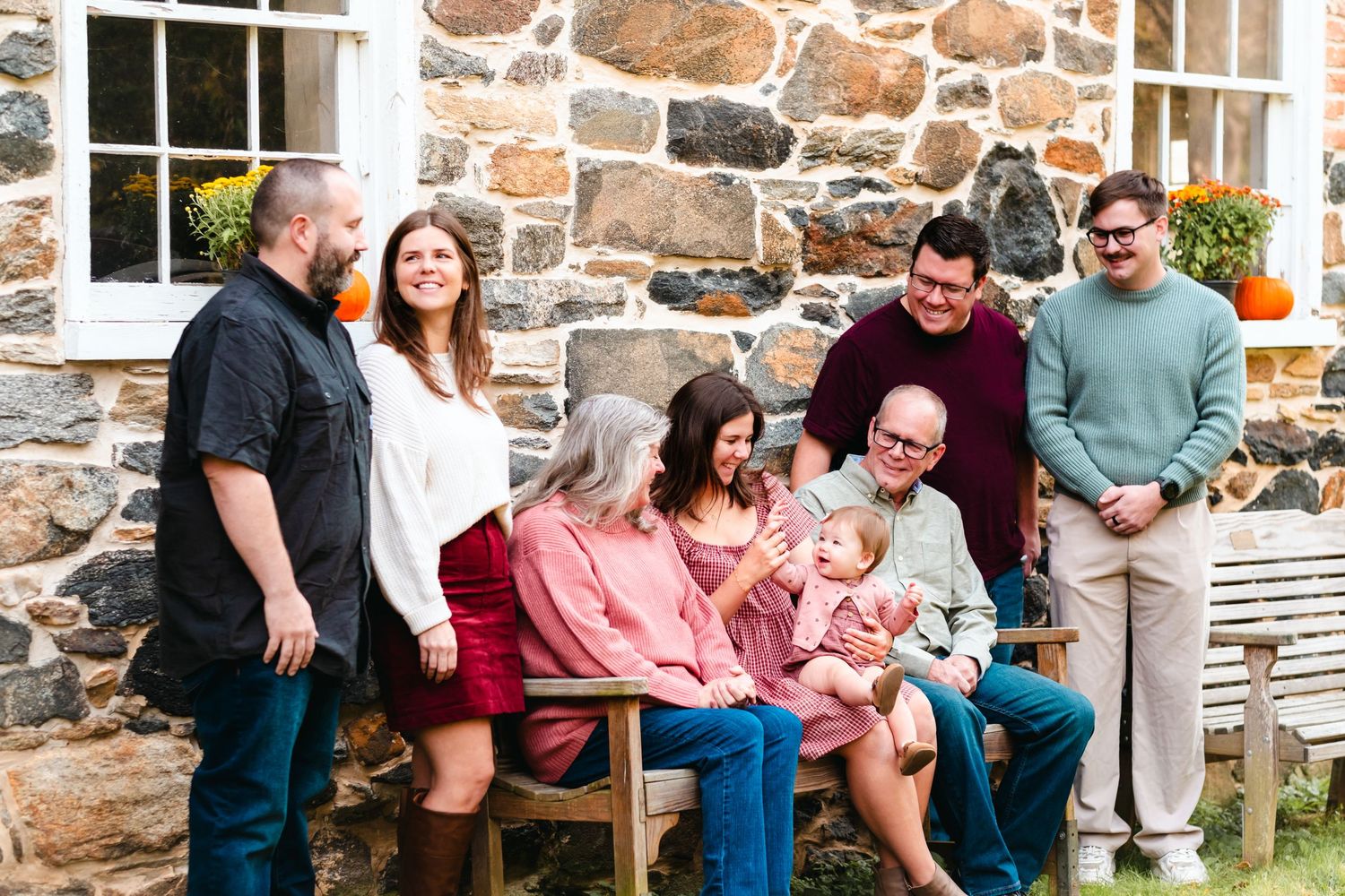 Multi-generational family gathering outdoors near stone building, sharing joyful moment with baby, pumpkins in background.