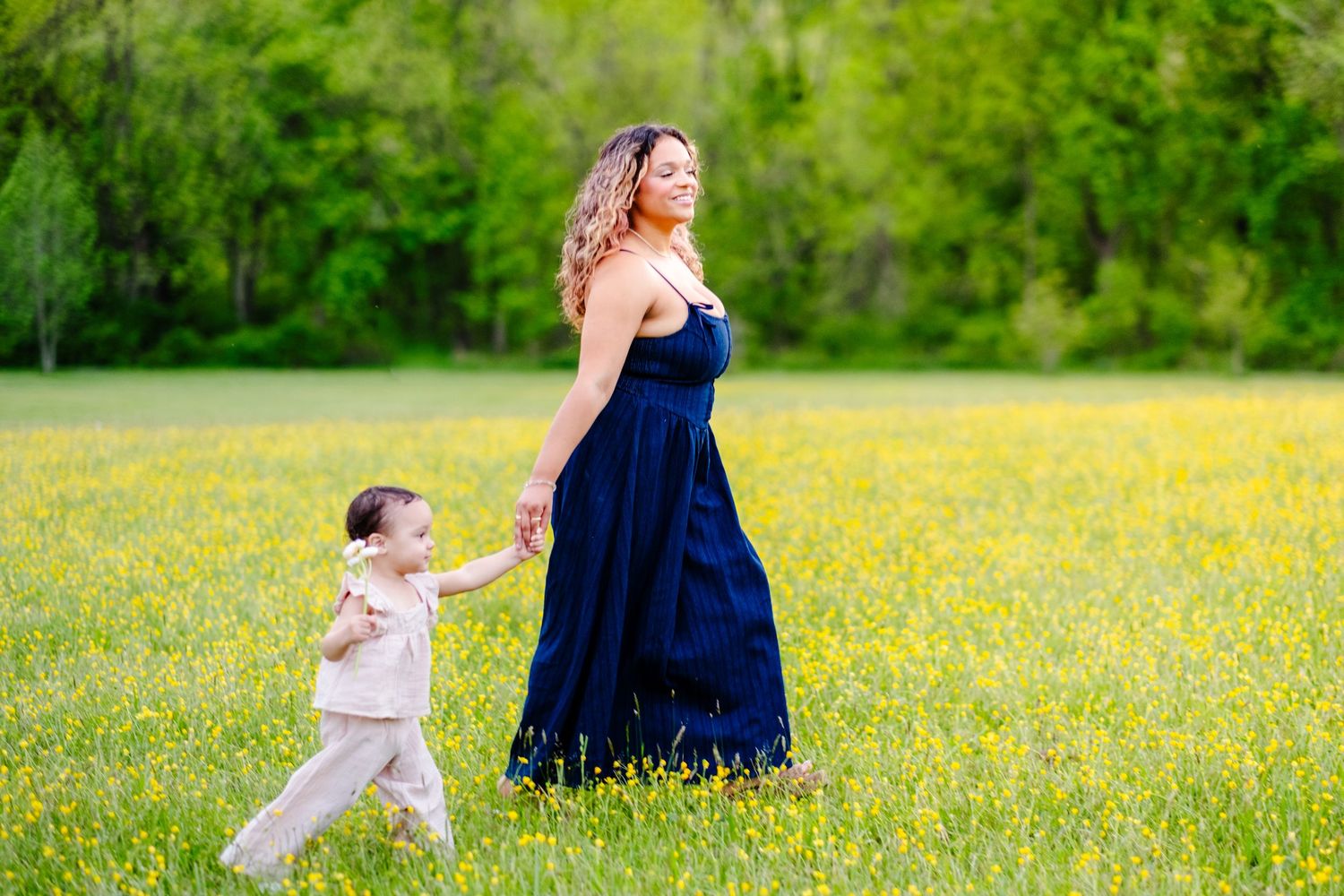 A mother in a navy dress holds her toddler's hand while walking through a sunny field of yellow wildflowers.