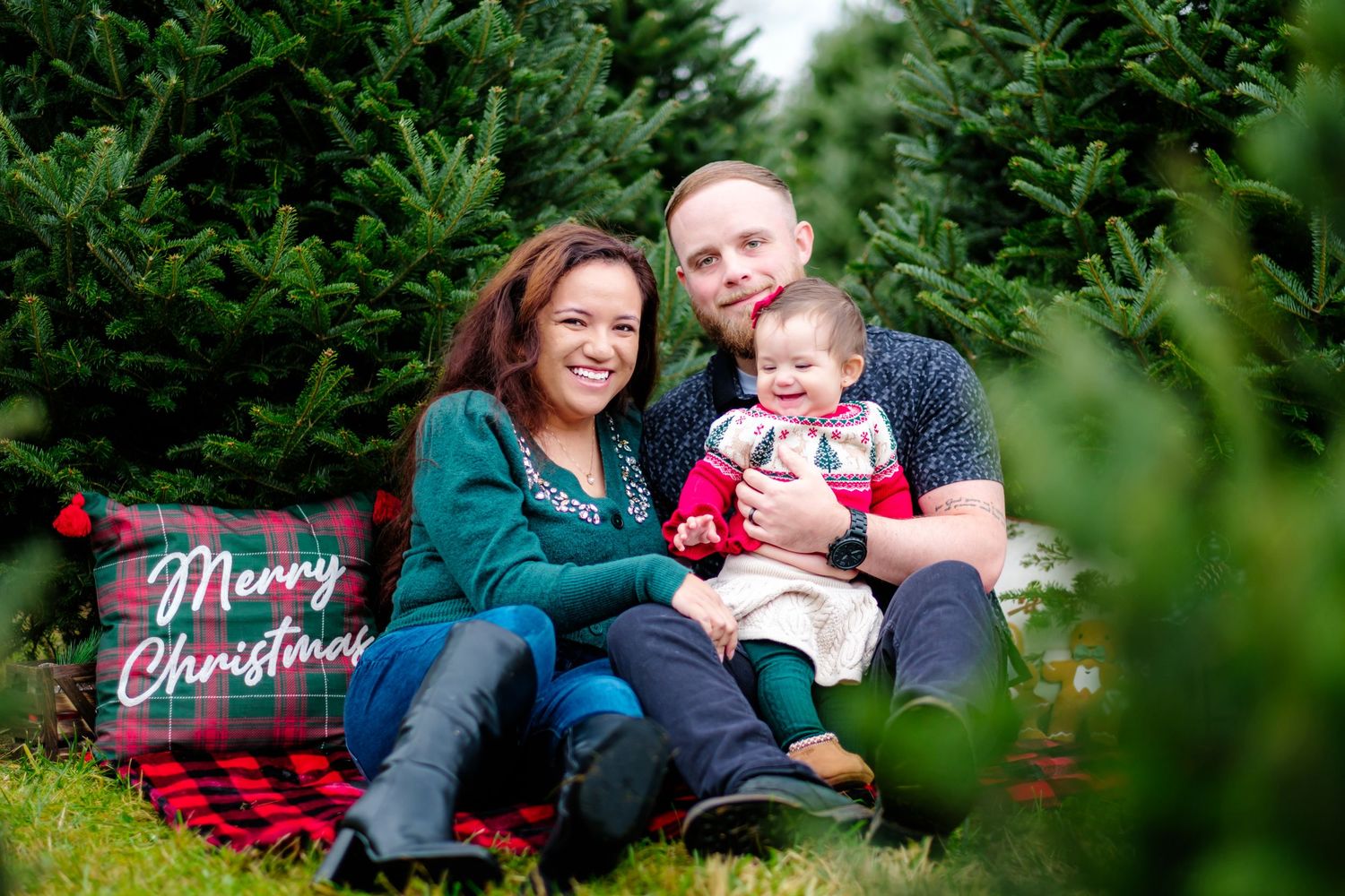 Family of three poses for Christmas photo at tree farm with Merry Christmas pillow, surrounded by lush green trees.